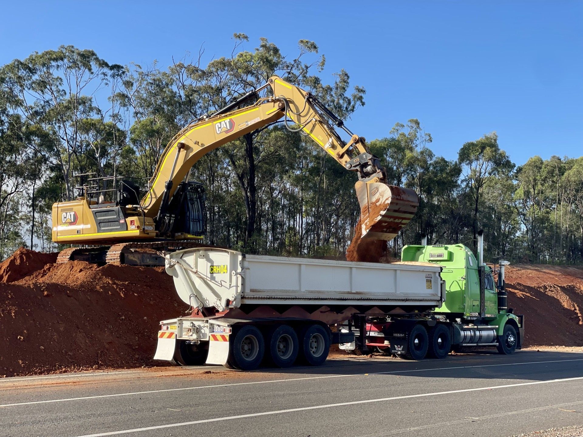 A yellow excavator loads red dirt into the back of a green semi-truck parked beside a road against a backdrop of trees. — LS Plant Hire in Calliope, QLD