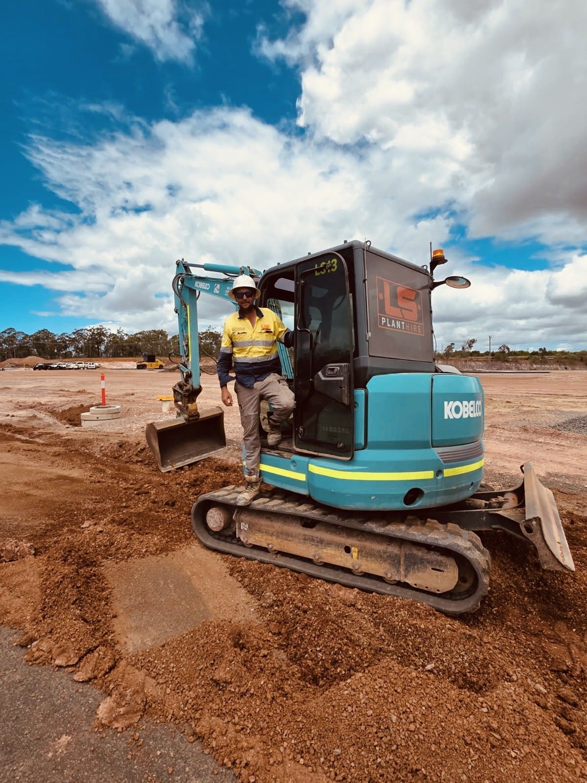 A person in high-visibility work gear stands in the cab of a teal excavator at an outdoor construction site. — LS Plant Hire in Calliope, QLD