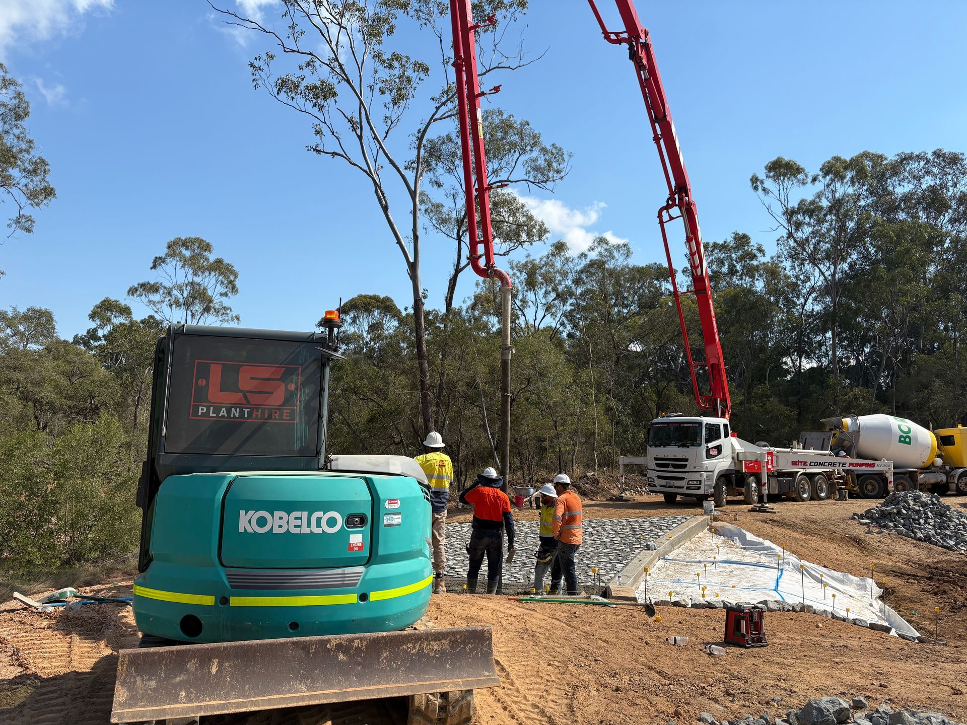 A green Kobelco excavator, workers, and a red concrete pump truck at a construction site with trees in the background. — LS Plant Hire in Calliope, QLD