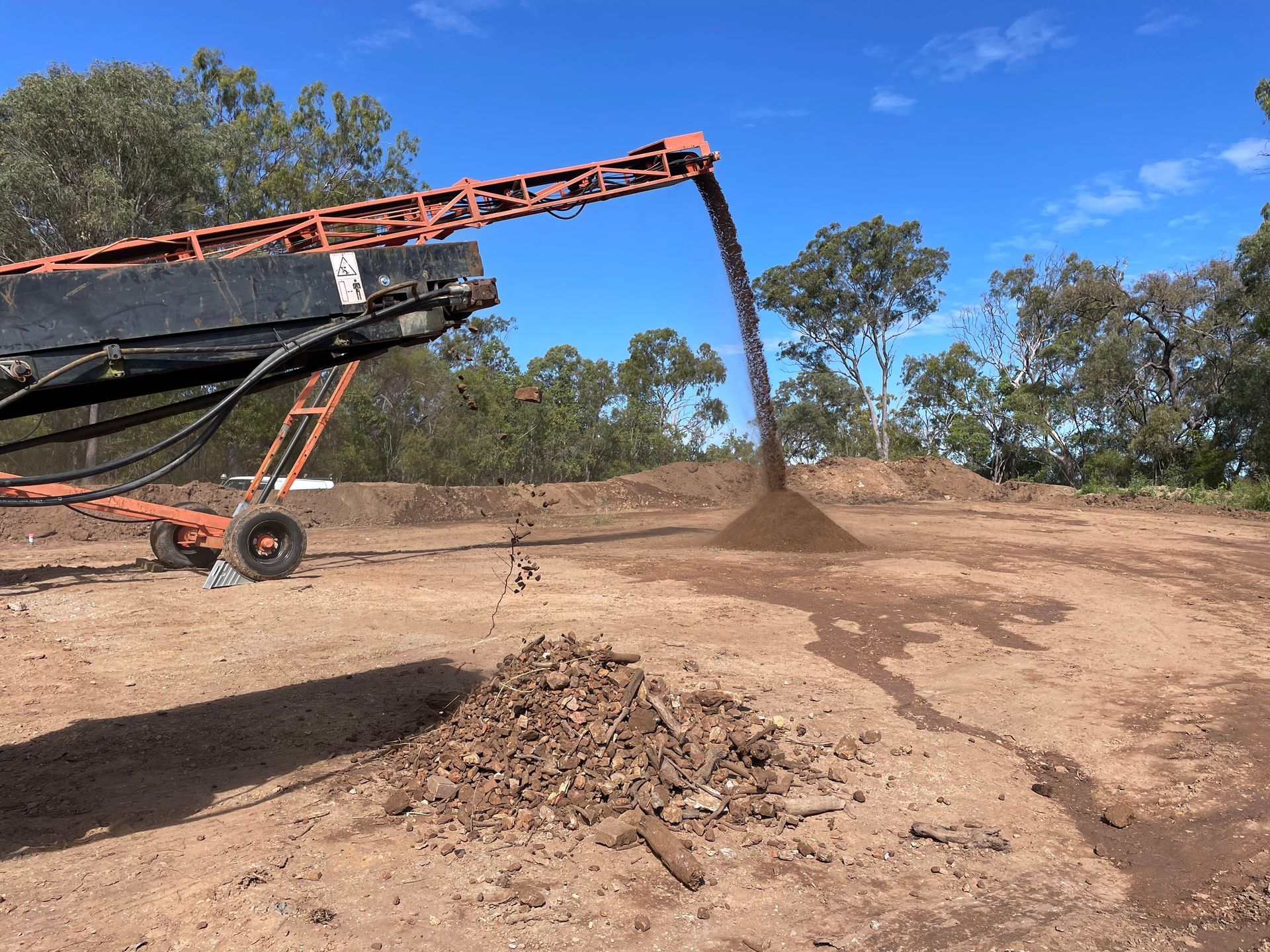 A conveyor belt machine depositing dirt and mulch into separate piles on a dirt field under a clear blue sky. — LS Plant Hire in Calliope, QLD