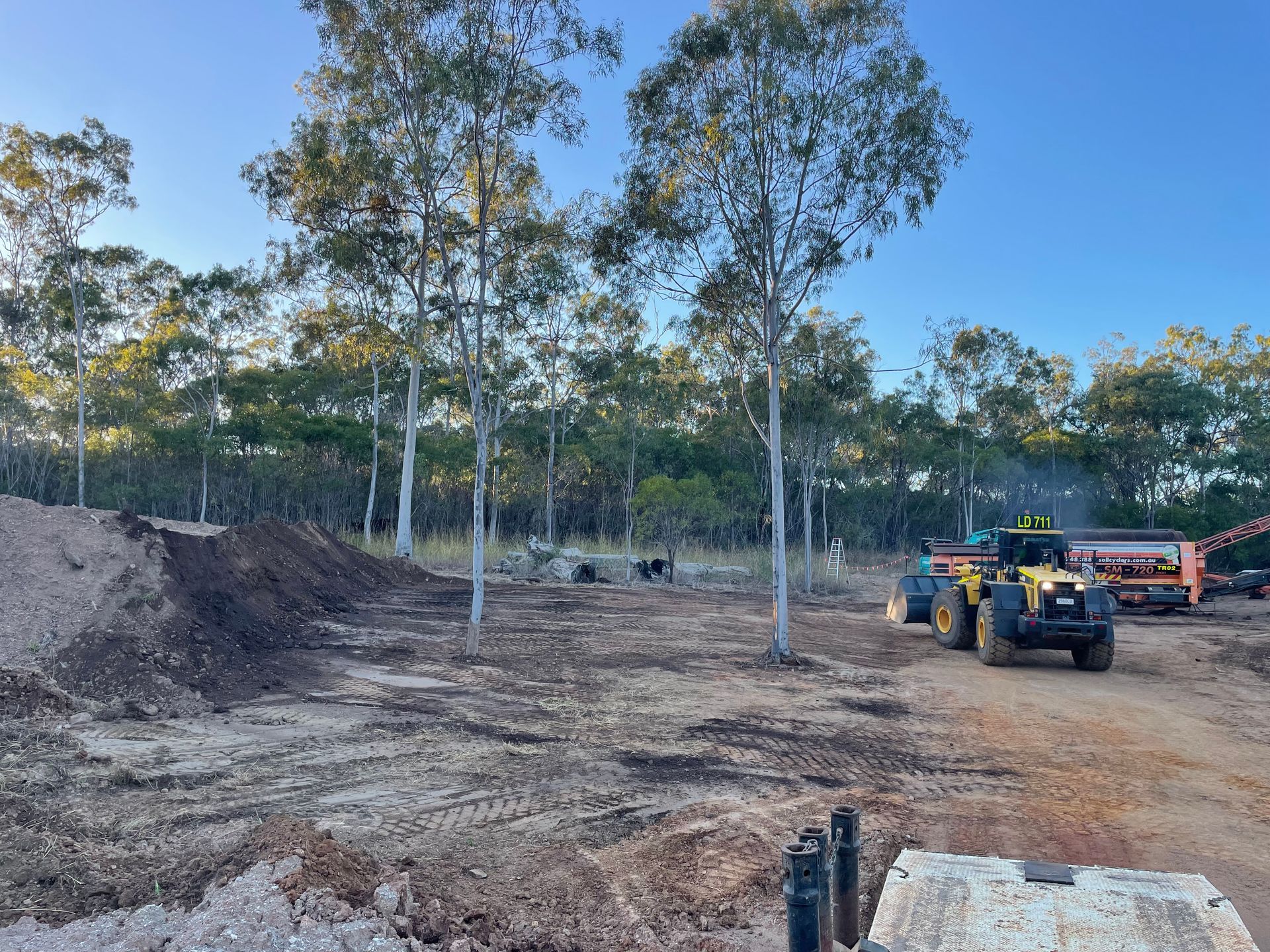A yellow construction tractor levels dirt on a cleared lot with trees under a clear blue sky. — LS Plant Hire in Calliope, QLD