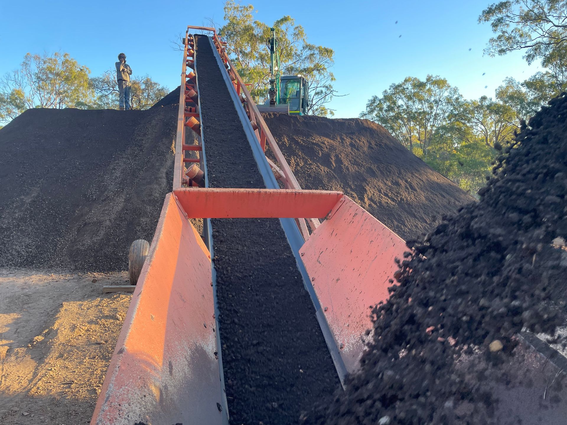 A conveyor belt transports dark mulch upward toward a pile outdoors, with a worker visible in the background. — LS Plant Hire in Calliope, QLD