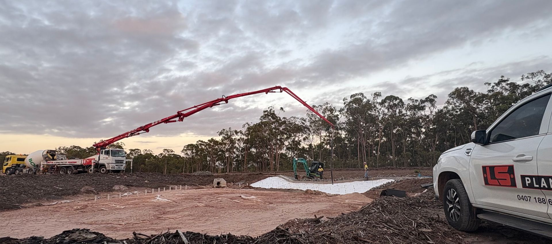 A concrete pump truck extends its boom to pour cement on a dirt construction site at sunset near a white pickup truck. — LS Plant Hire in Calliope, QLD