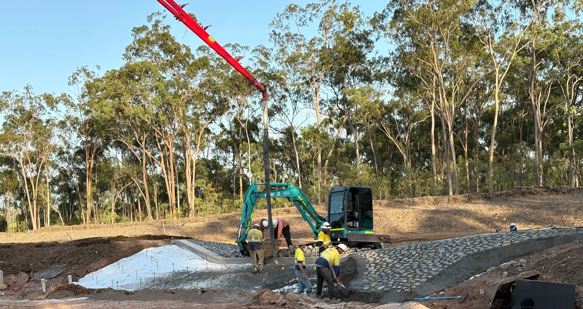 Construction workers use a pump truck and excavator to pour concrete onto a rock-filled area at a wooded job site. — LS Plant Hire in Calliope, QLD