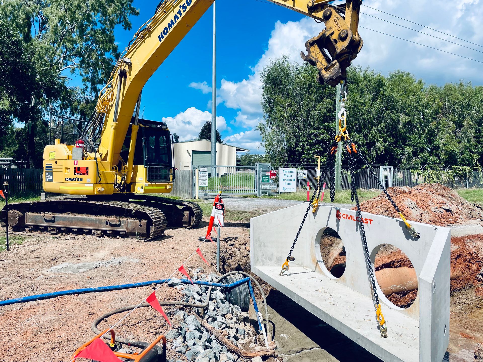 A yellow excavator lifts a heavy, L-shaped precast concrete headwall structure over a dirt construction site. — LS Plant Hire in Calliope, QLD