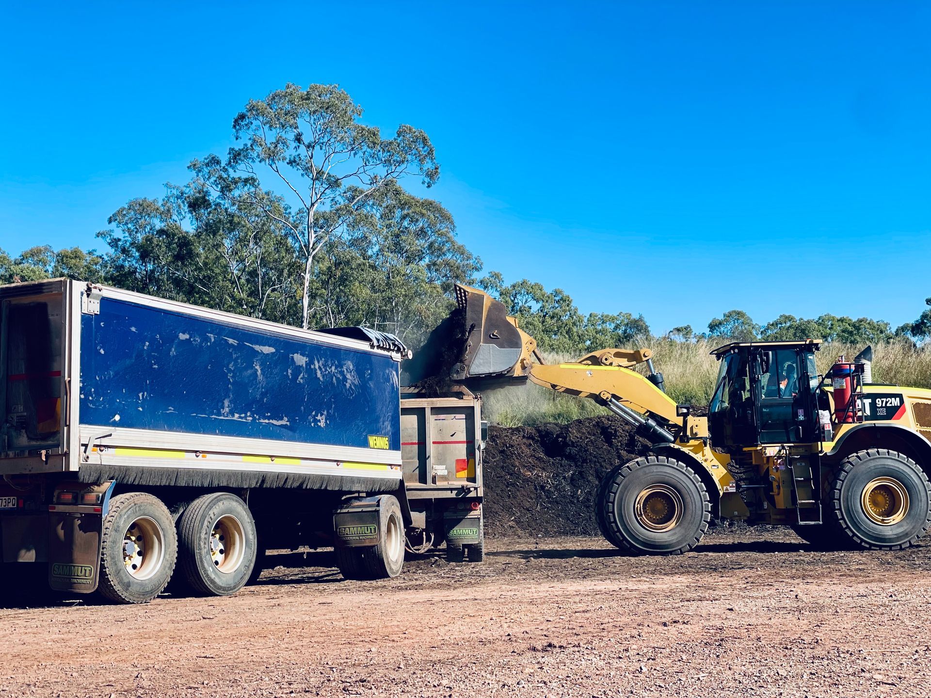 A yellow front-end loader dumps dirt into a large, blue-and-white dump truck parked on a dirt lot under a clear blue sky. — LS Plant Hire in Calliope, QLD