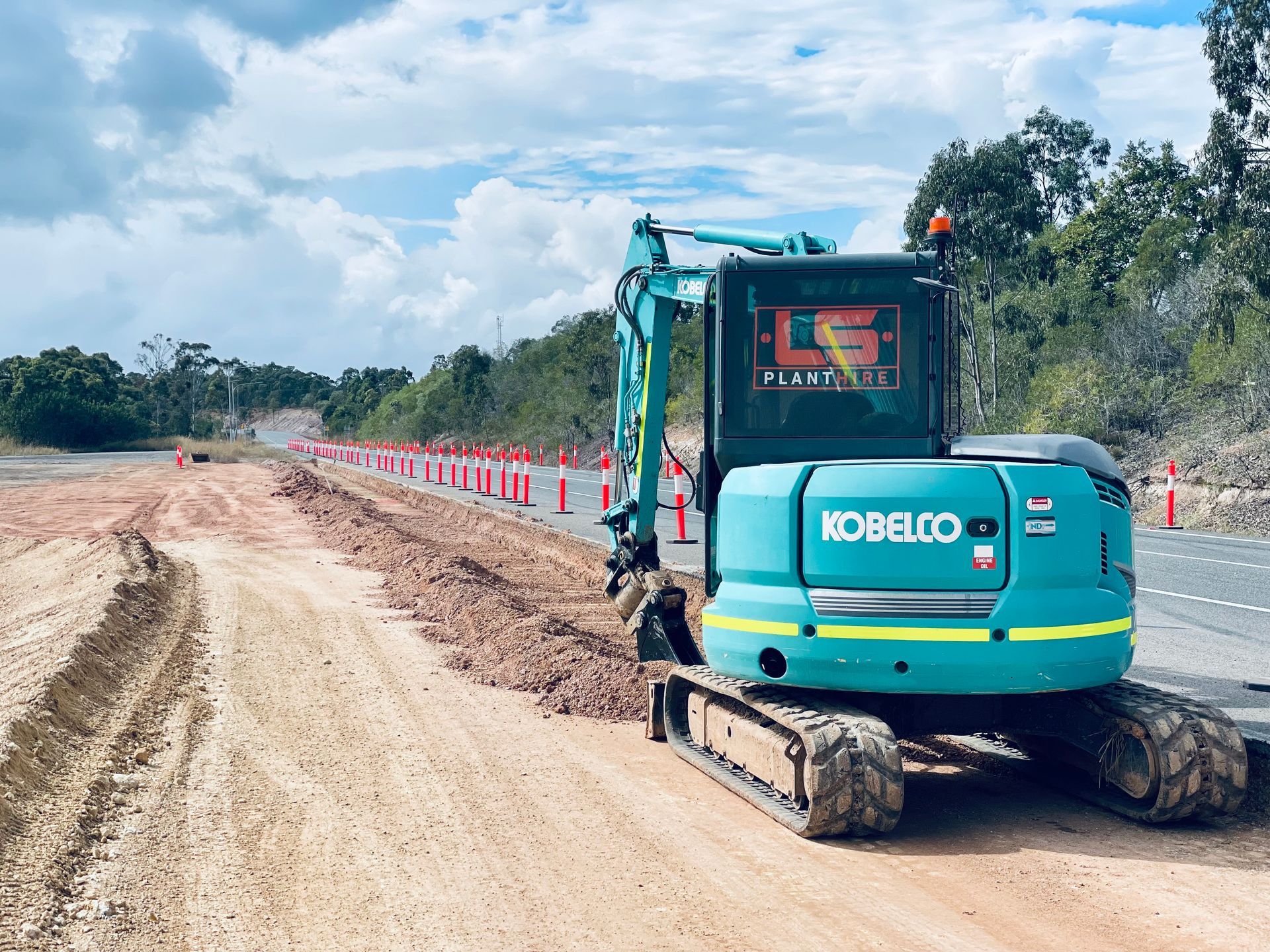 A teal Kobelco excavator sits on a dirt shoulder next to a paved road lined with orange safety cones under a cloudy sky. — LS Plant Hire in Calliope, QLD