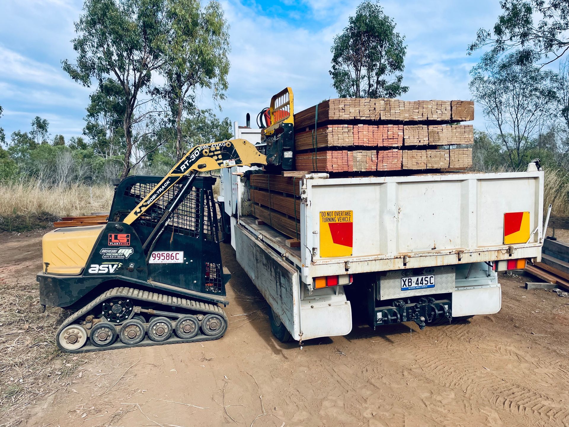 A black tracked skid steer loader loads a stack of timber beams into the white tray of a truck on a dirt road. — LS Plant Hire in Calliope, QLD