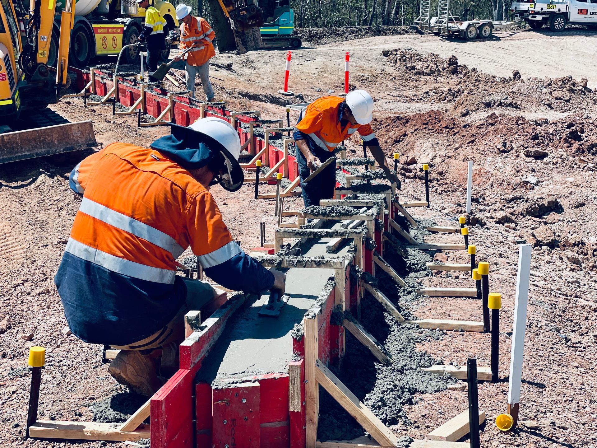 Construction workers in orange high-visibility clothing smooth fresh concrete into red wooden forms at a job site. — LS Plant Hire in Calliope, QLD