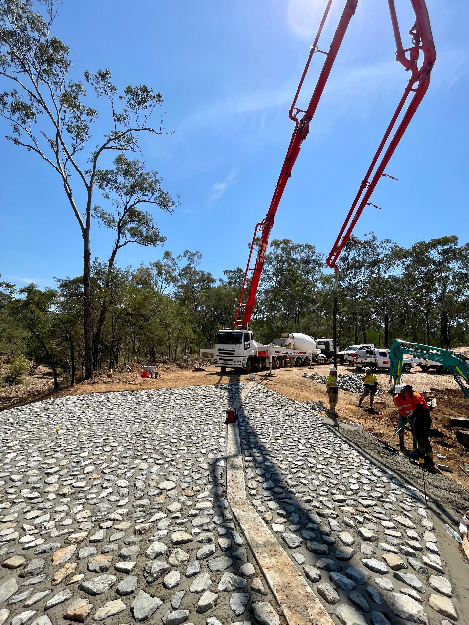 A red concrete pump truck pours concrete over a rock-lined area at a sunny outdoor construction site with workers nearby. — LS Plant Hire in Calliope, QLD