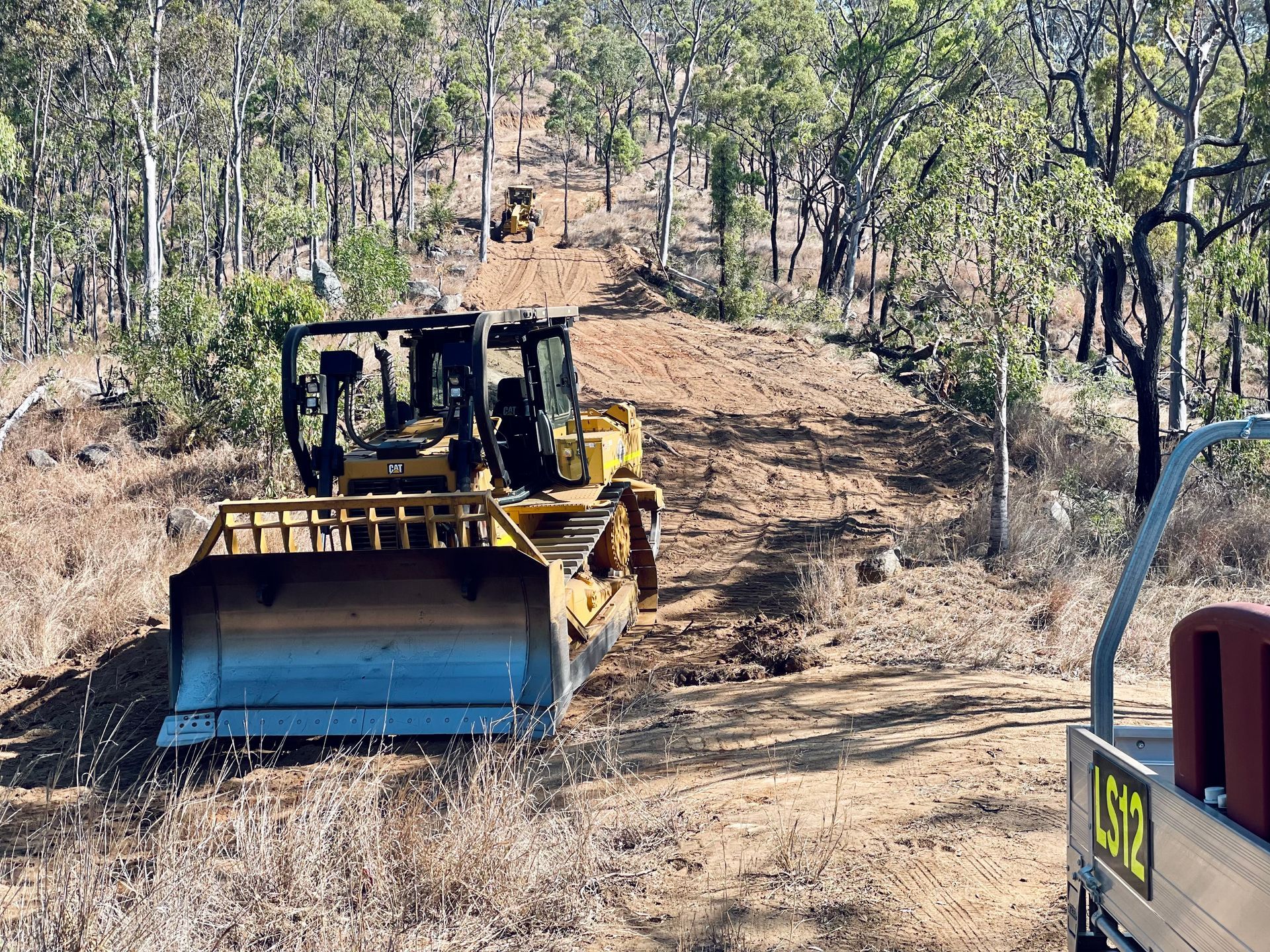 A yellow bulldozer clears a path through a sunlit, forested area, with a second piece of machinery visible further ahead. — LS Plant Hire in Calliope, QLD