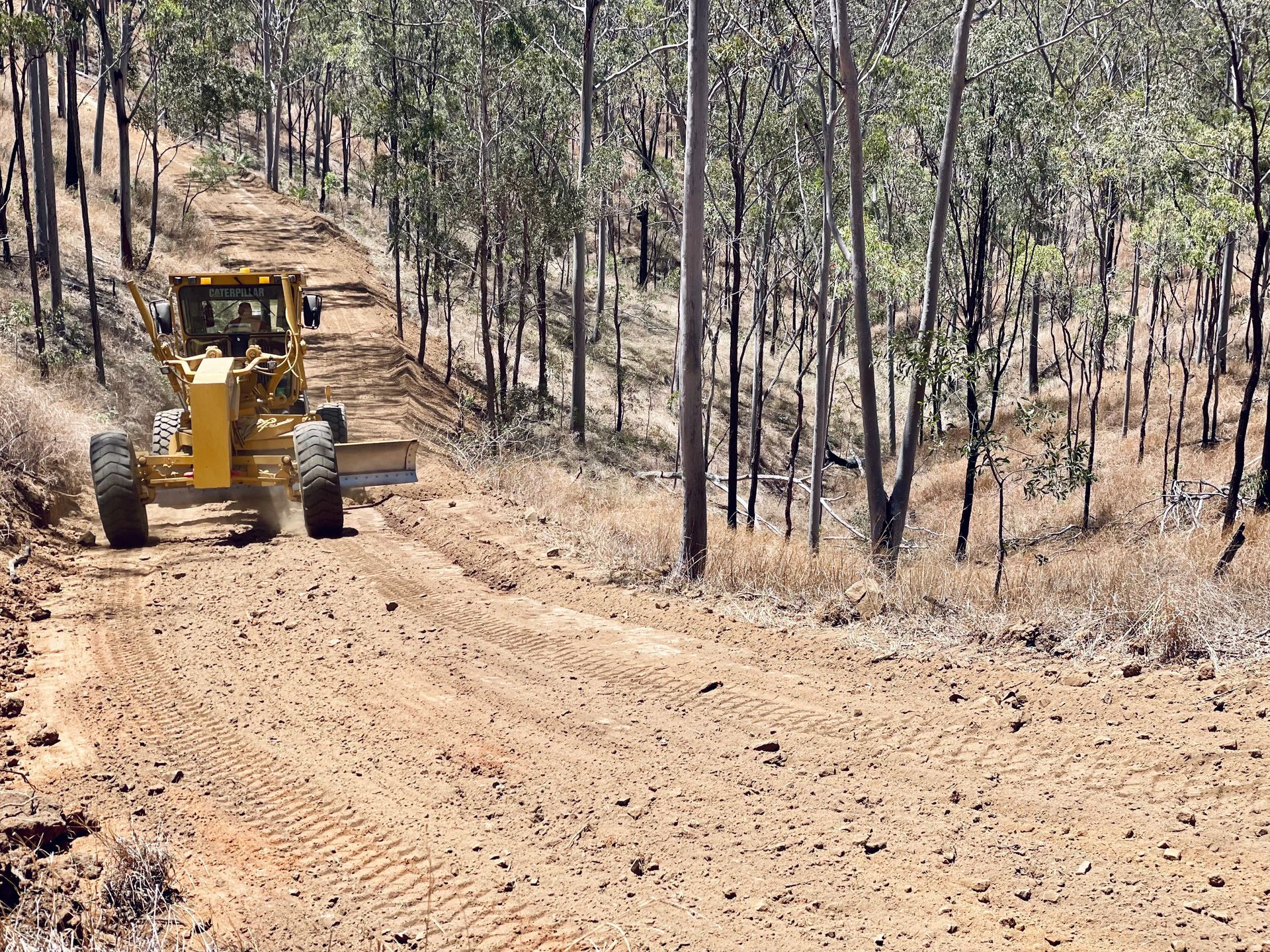 A yellow motor grader smoothing a dirt road through a wooded area on a sunny day. — LS Plant Hire in Calliope, QLD