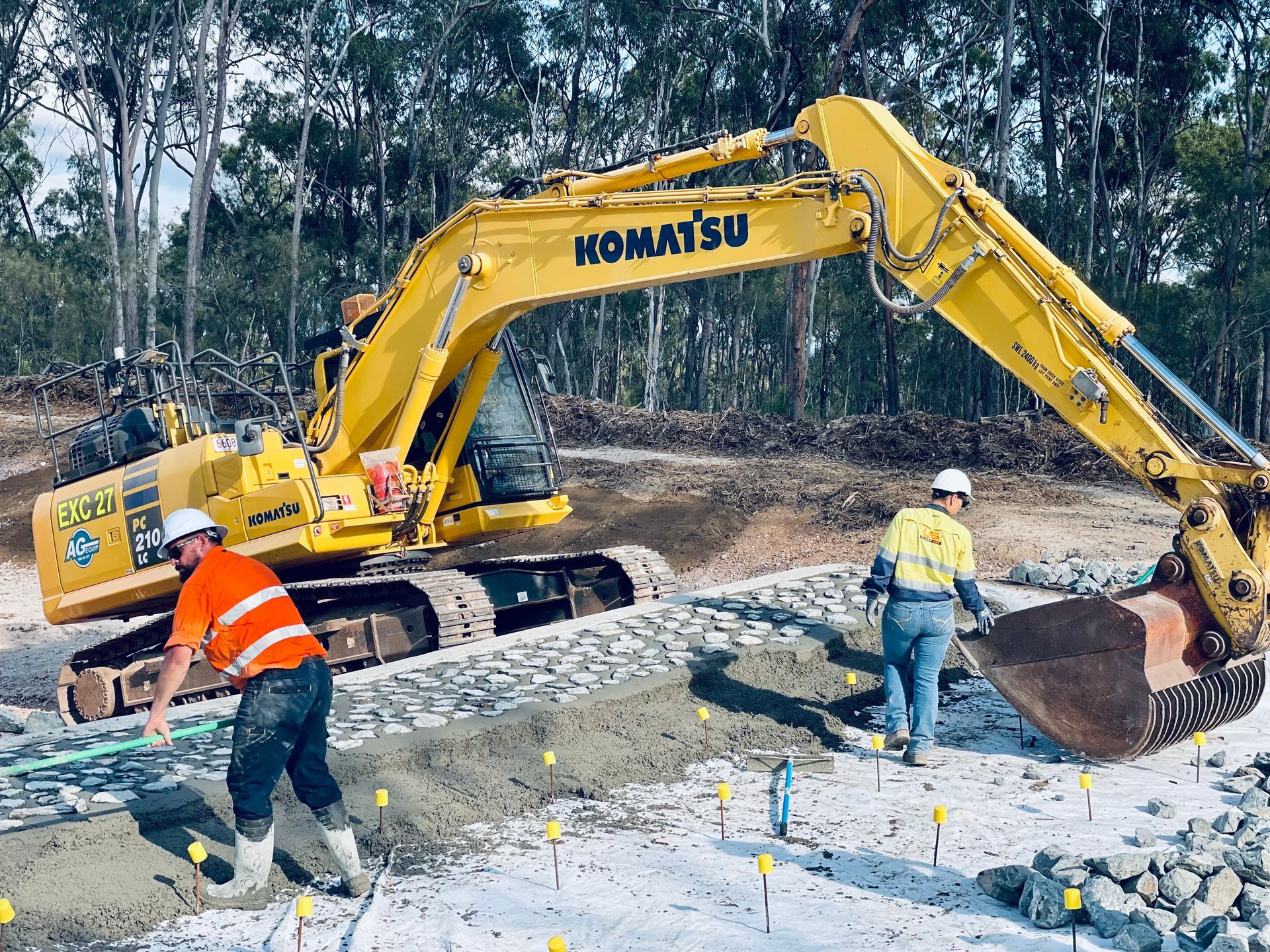 A yellow Komatsu excavator works on a dirt construction site with two workers in high-visibility vests nearby. — LS Plant Hire in Calliope, QLD