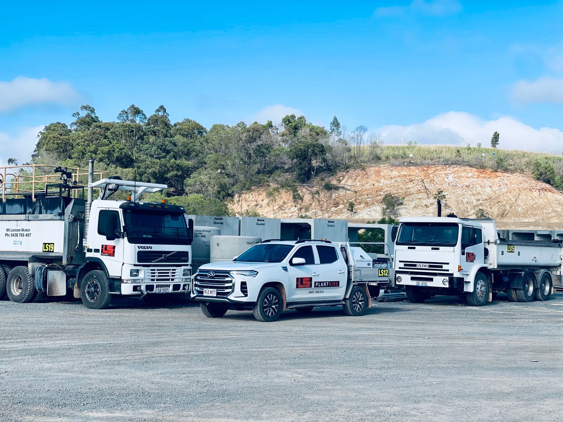 A white pickup truck parked between two heavy-duty industrial trucks at a gravel site under a blue sky. — LS Plant Hire in Calliope, QLD