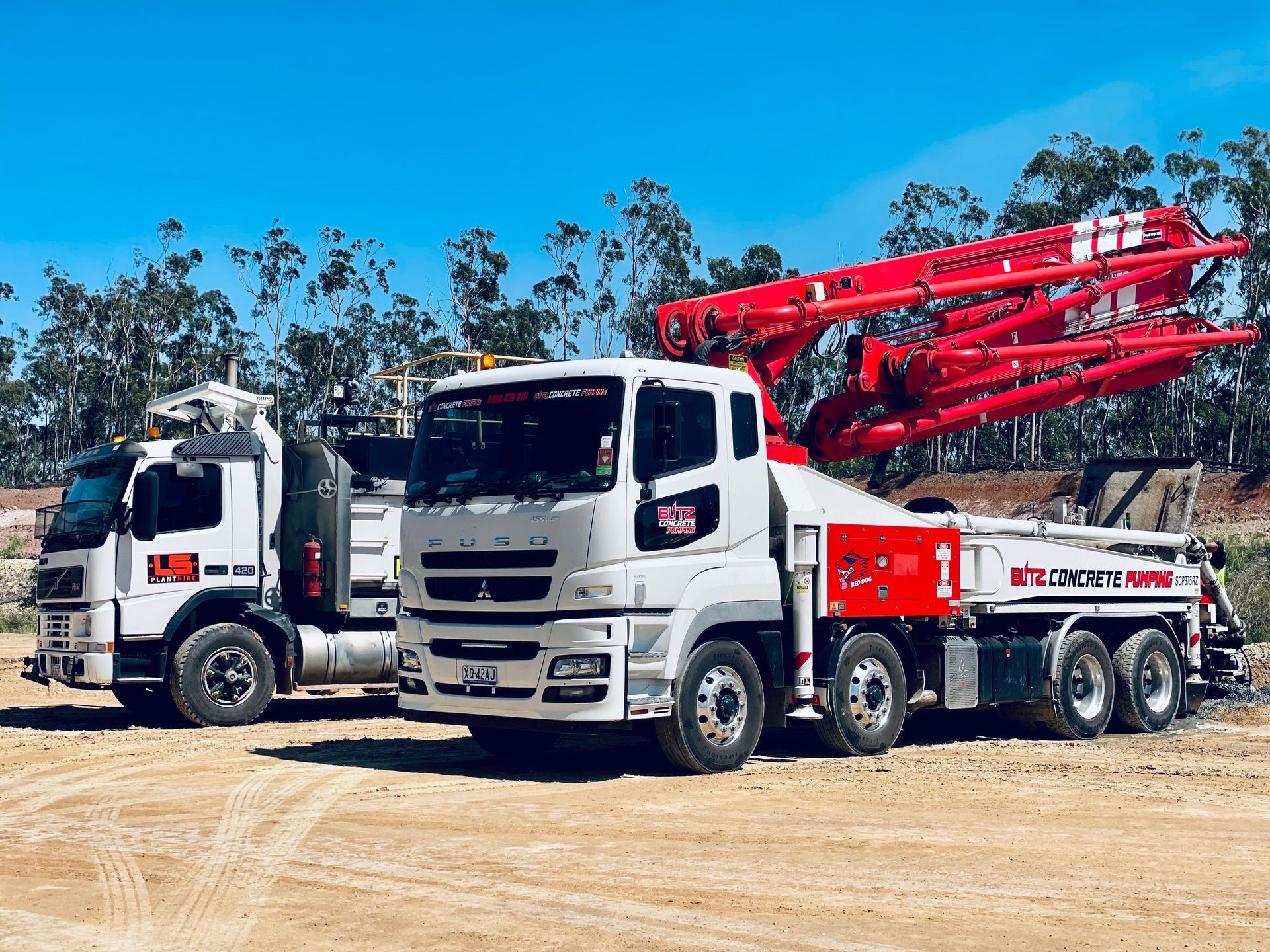 Two white concrete pump trucks with red booms parked on a dirt lot against a clear blue sky. — LS Plant Hire in Calliope, QLD