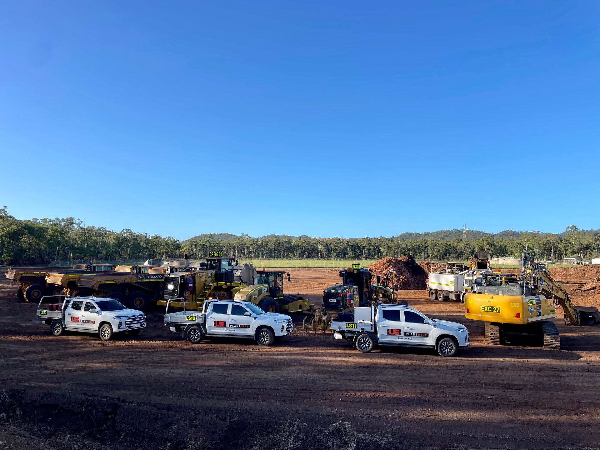 A line of three white service trucks parked on a dirt construction site with yellow heavy machinery under a blue sky. — LS Plant Hire in Calliope, QLD