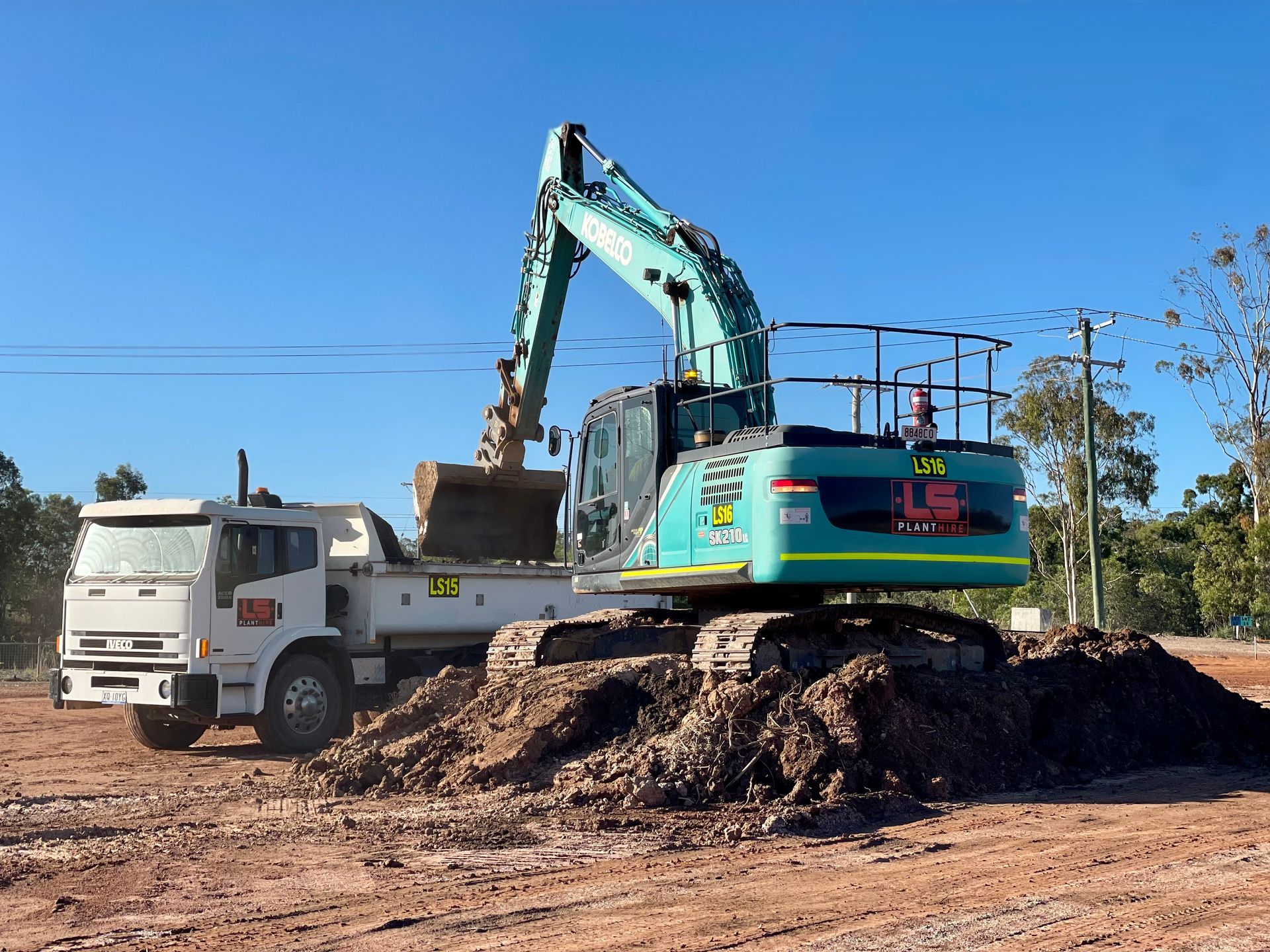 A teal excavator loading soil into a white dump truck at a dirt construction site under a clear blue sky. — LS Plant Hire in Calliope, QLD