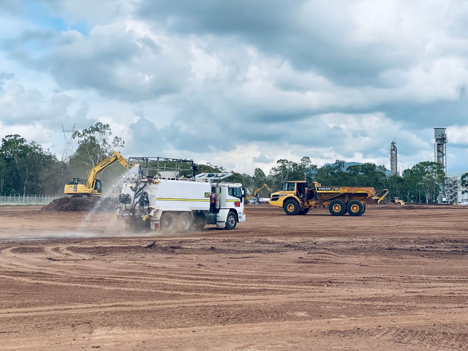A white water truck sprays dust on a dirt construction site, with an excavator and a dump truck working in the background. — LS Plant Hire in Calliope, QLD