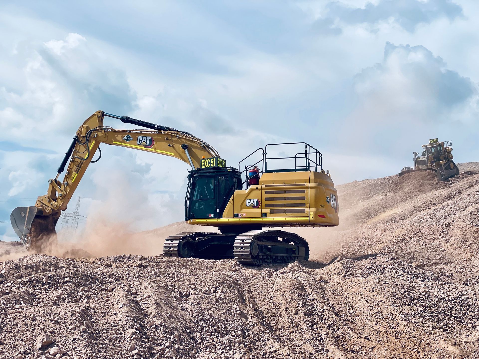 A yellow Caterpillar excavator operates on a dusty, hilly construction site under a cloudy blue sky. — LS Plant Hire in Calliope, QLD