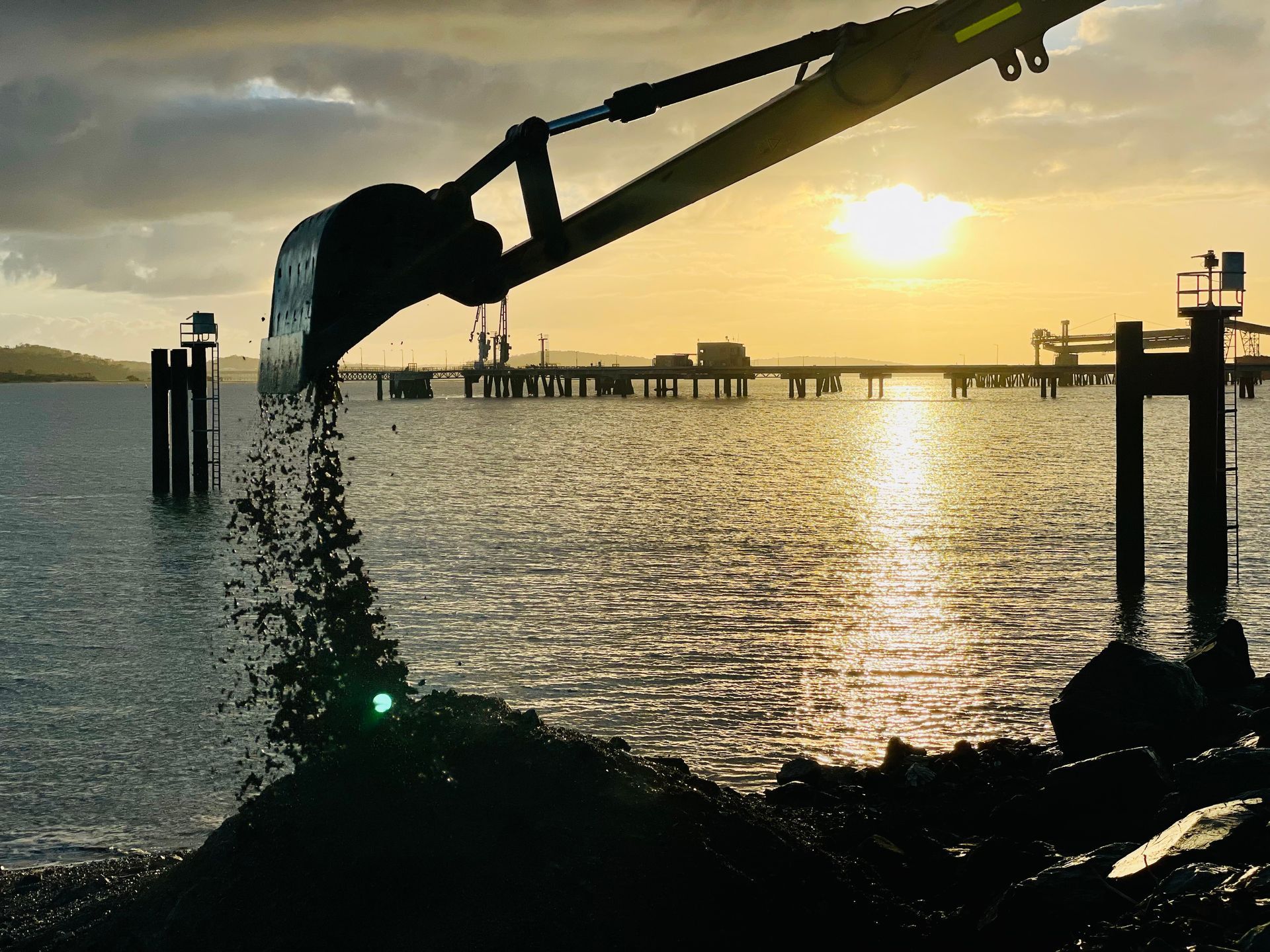 An excavator bucket pouring sand into water at sunset near a pier. — LS Plant Hire in Calliope, QLD