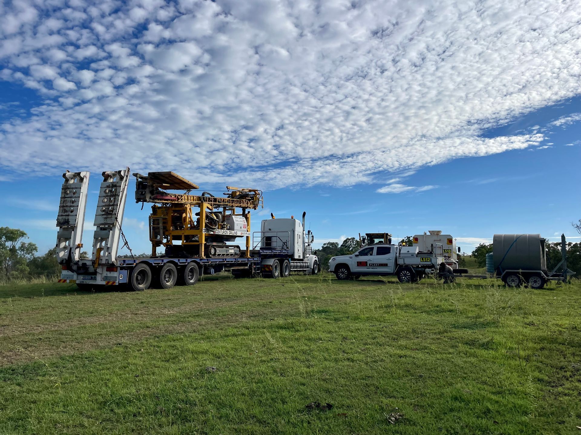 A semi-truck carrying a large yellow drilling rig parked in a grassy field under a bright, cloudy blue sky. — LS Plant Hire in Calliope, QLD