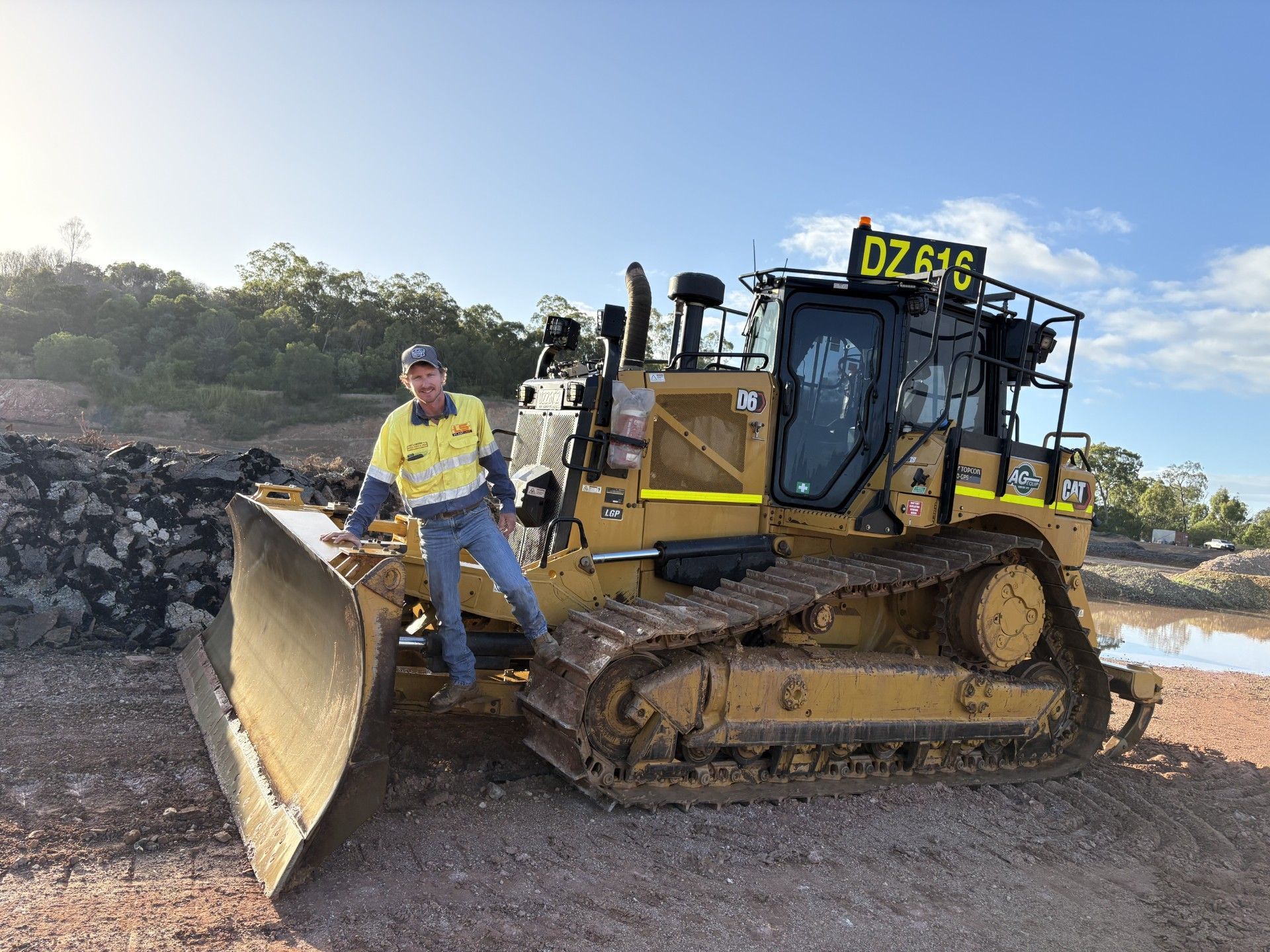 A construction worker in a high-visibility uniform stands on the blade of a large yellow bulldozer marked DZ615 outdoors. — LS Plant Hire in Calliope, QLD