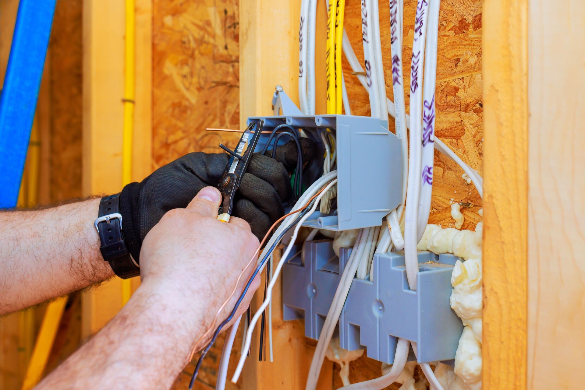 Electrician wearing black gloves strips insulation from electrical wires inside a plastic junction box on a wood frame wall.