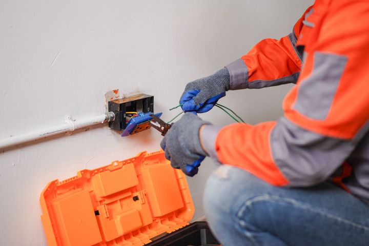 An electrician in an orange high-visibility jacket uses pliers to strip green electrical wires at a wall-mounted outlet.