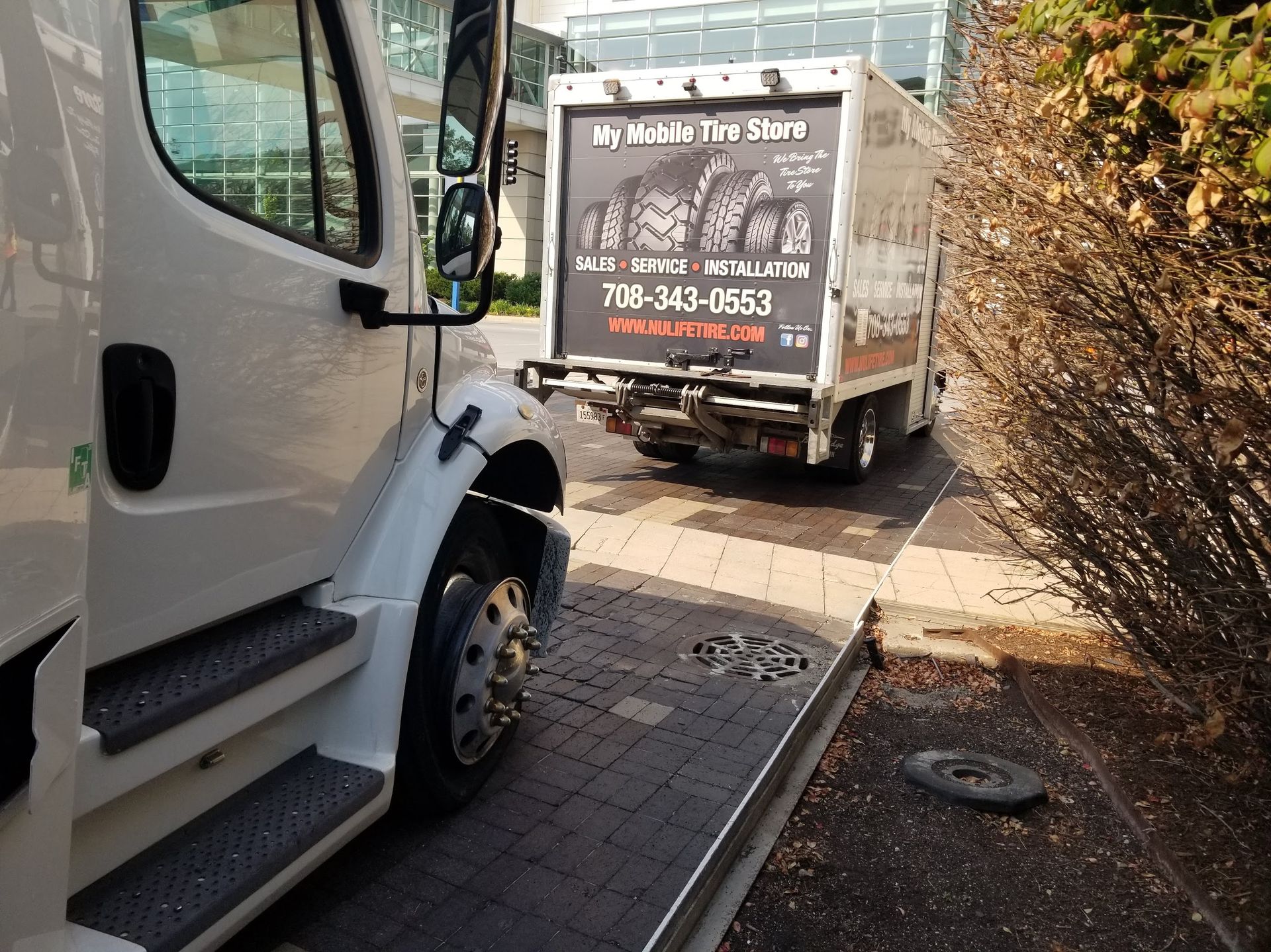 White truck towing a service truck with tire shop advertisement in front of building.