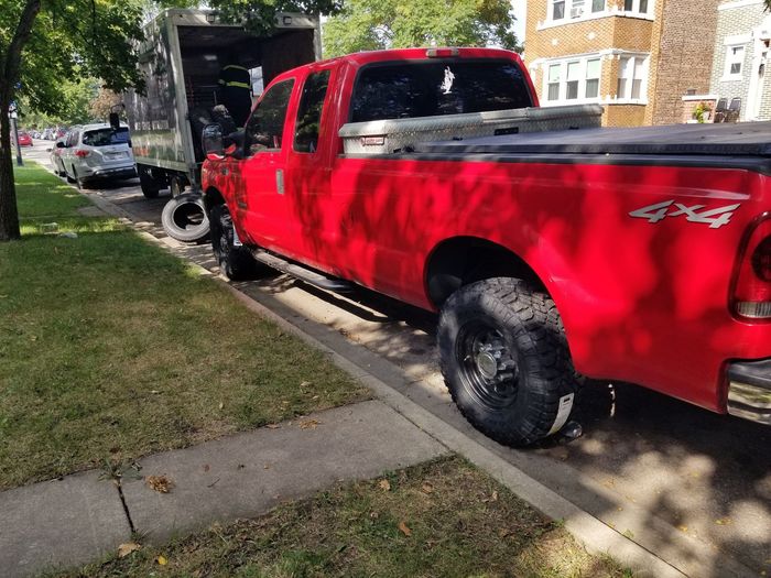 Red pickup truck parked next to a curb; moving truck in background, and a car.