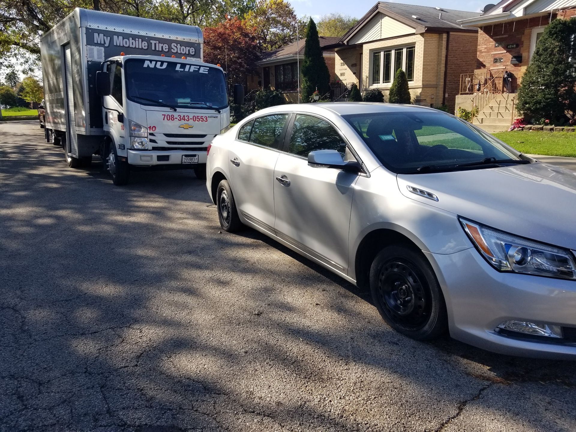 Moving truck and silver car parked on a street in front of houses on a sunny day.