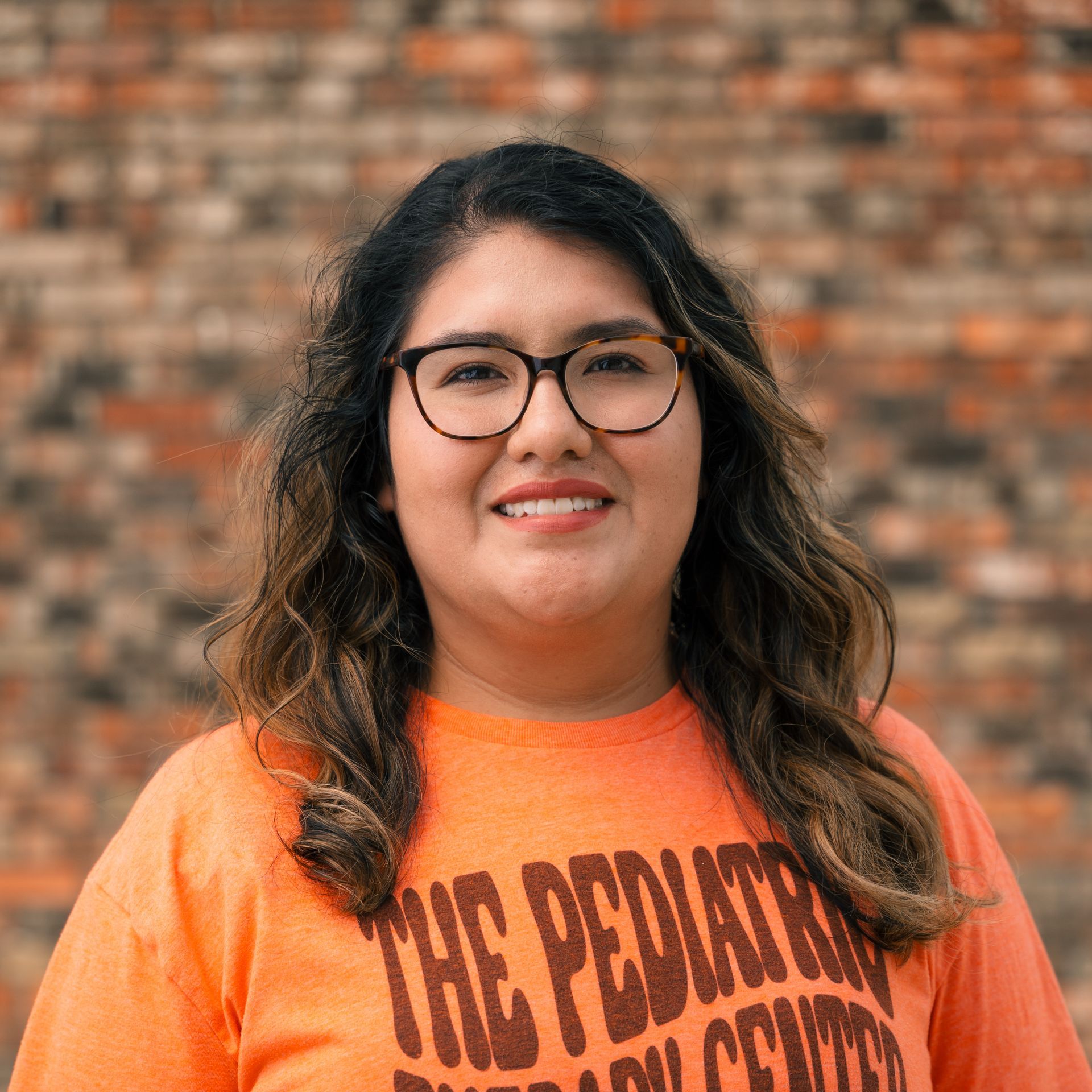 A woman wearing glasses and an orange shirt is standing in front of a brick wall.