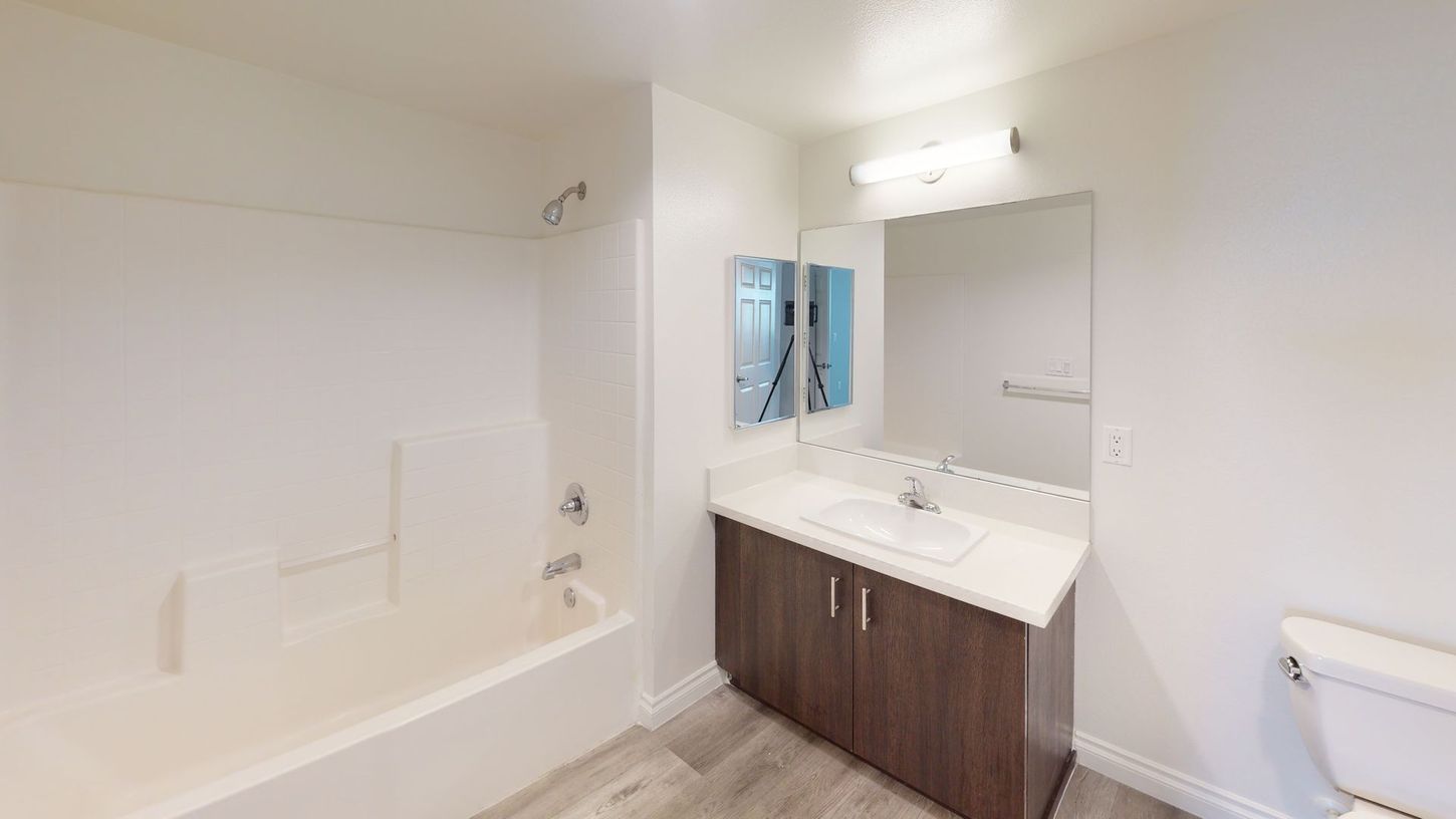 Bathroom with white tub, brown vanity, mirror, and white toilet. Gray floors, white walls.