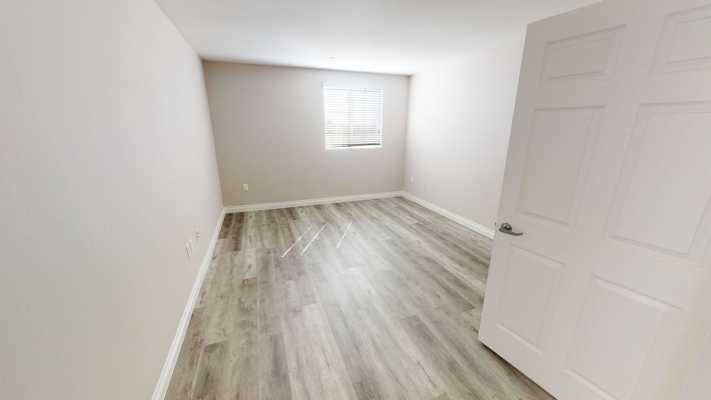 Empty room with gray-toned wood flooring, light walls, a window, and a closed white door.