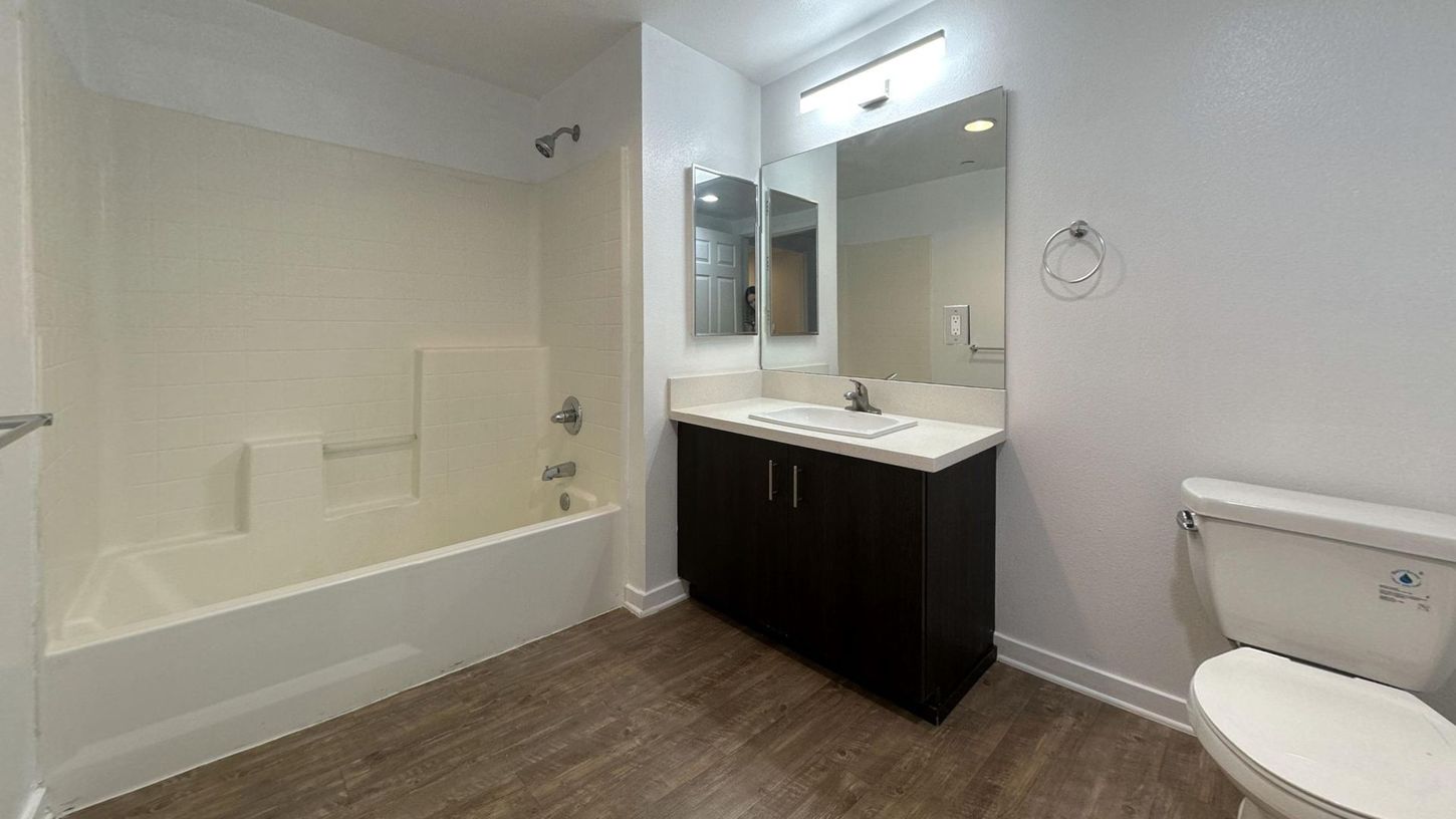 Bathroom with white tub/shower, dark vanity, large mirror, and toilet, with wood-look flooring.
