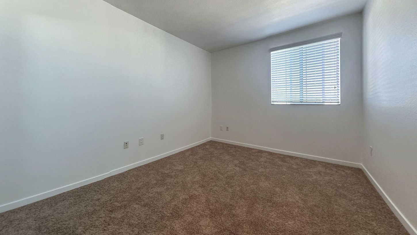 Empty room with white walls, brown carpet, window with blinds.