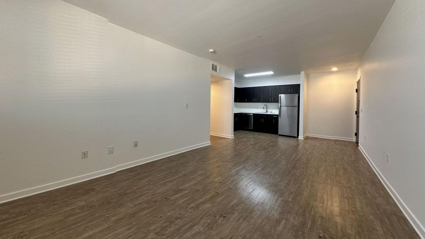 Empty living space with laminate flooring, white walls, and a glimpse of a kitchen.