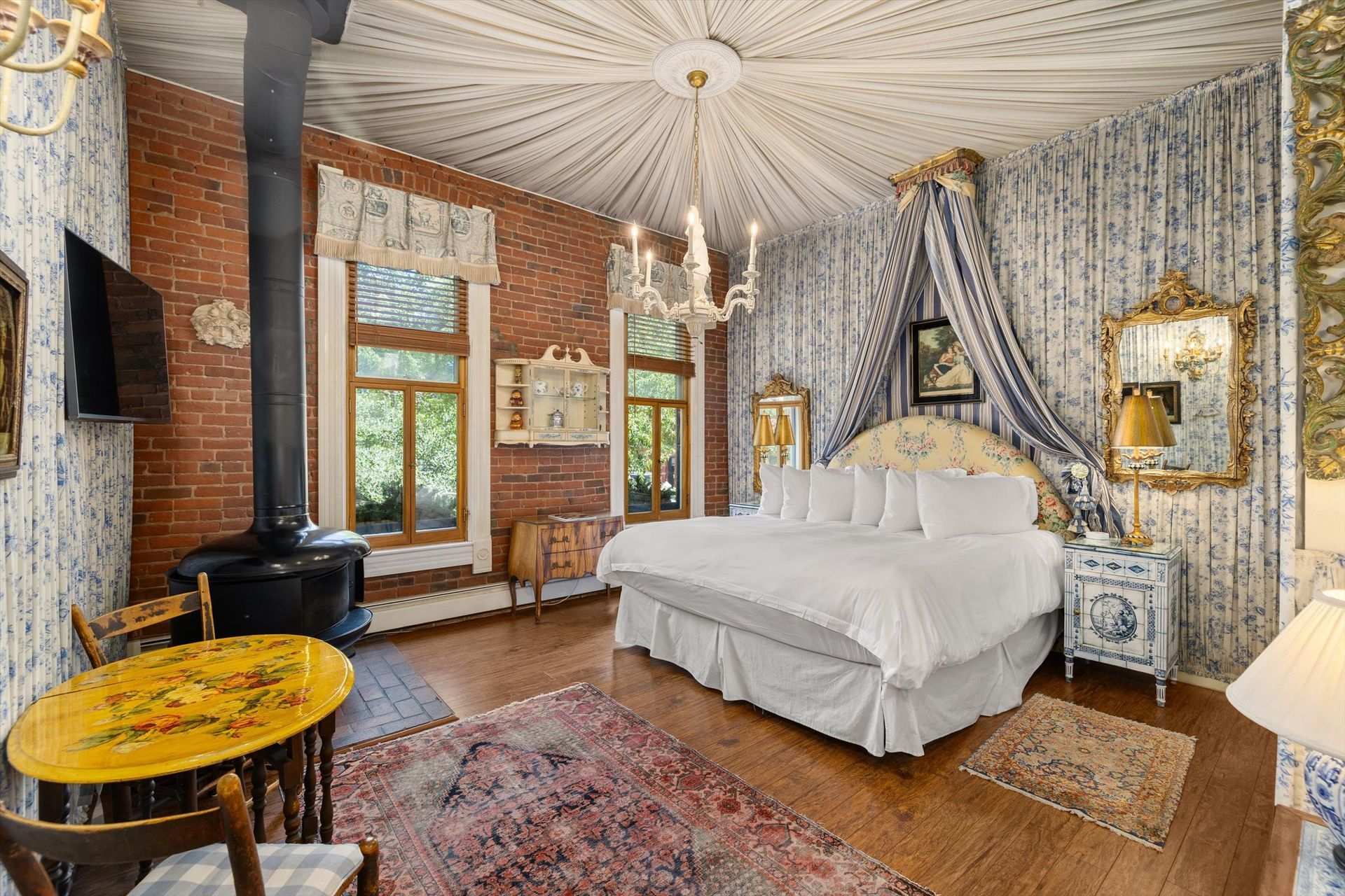 Bedroom with exposed brick, blue floral wallpaper, chandelier, canopy bed, wood-burning stove, and ornate furniture.