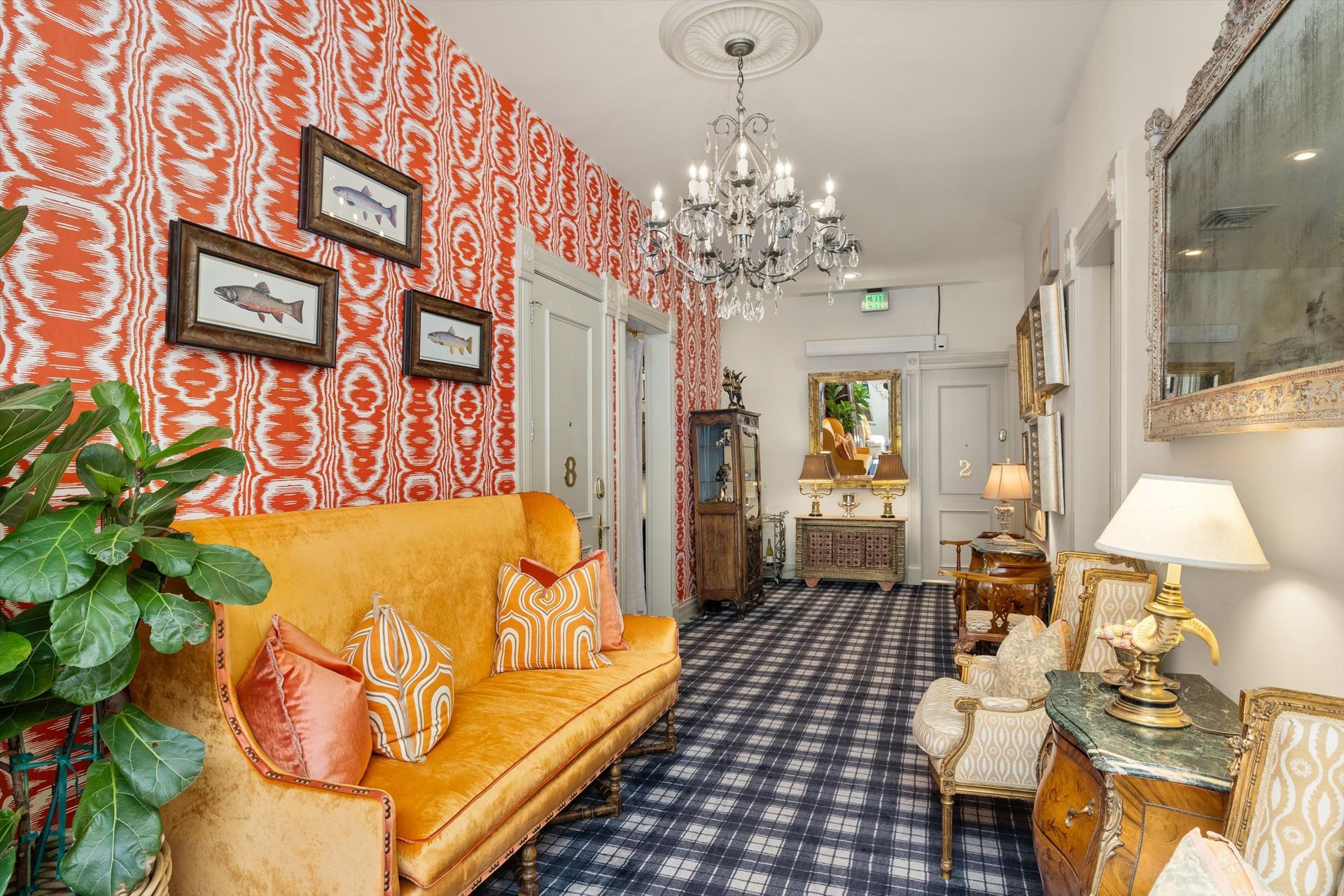 A hallway with orange patterned wallpaper, an orange couch, and decorative furniture. A chandelier hangs from the ceiling.