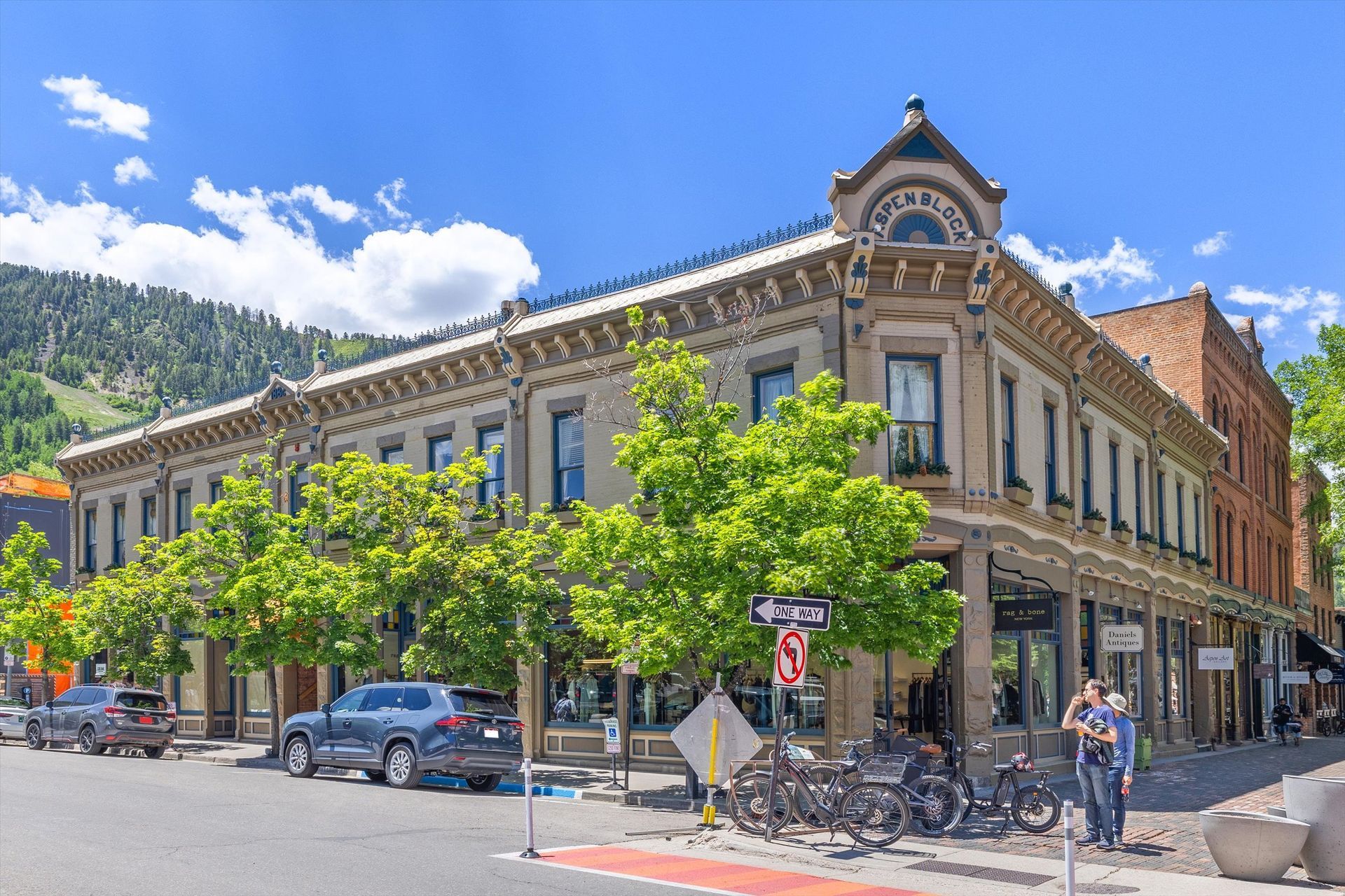 Historic brick buildings on a street corner in a mountain town; trees line the sidewalk, and a mountain is in the background.