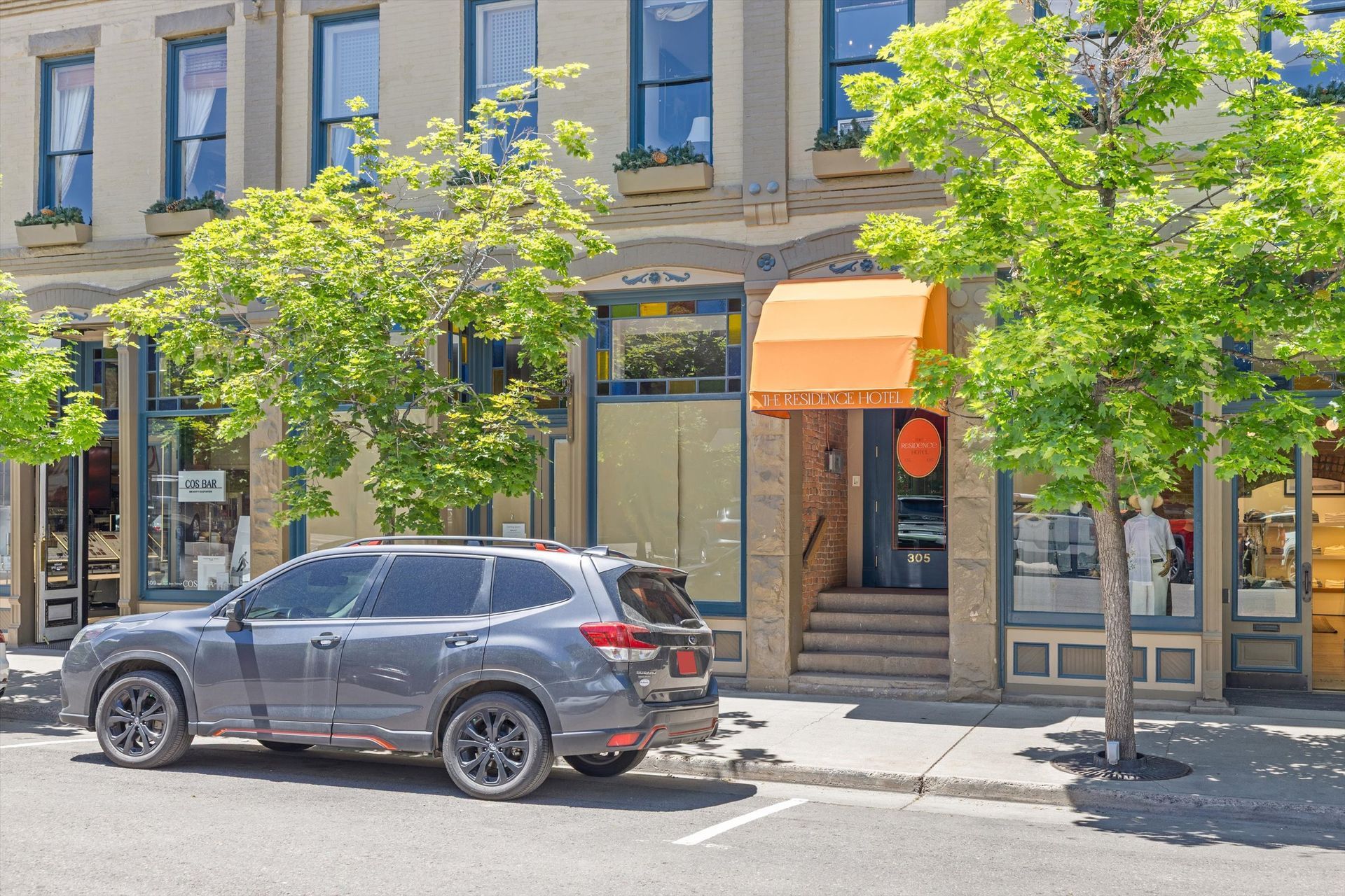 A gray SUV parked in front The Residence Hotel with a bright orange awning and several businesses on a sunny day.