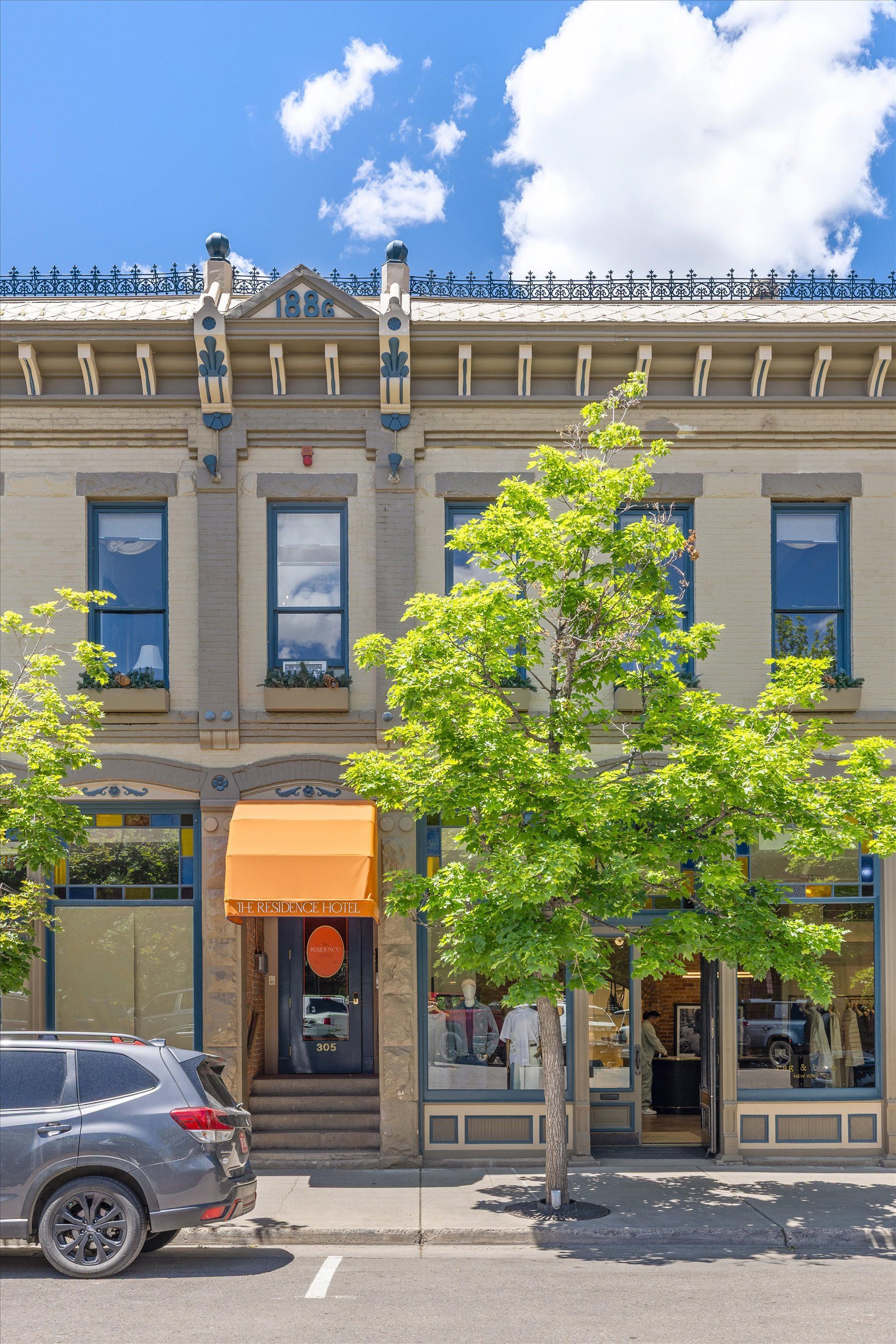 A two-story building, which is  The Residence Hotel, with shops, a tan facade, blue-framed windows, and an orange awning. A tree stands in front, and a dark gray car is parked on the street.