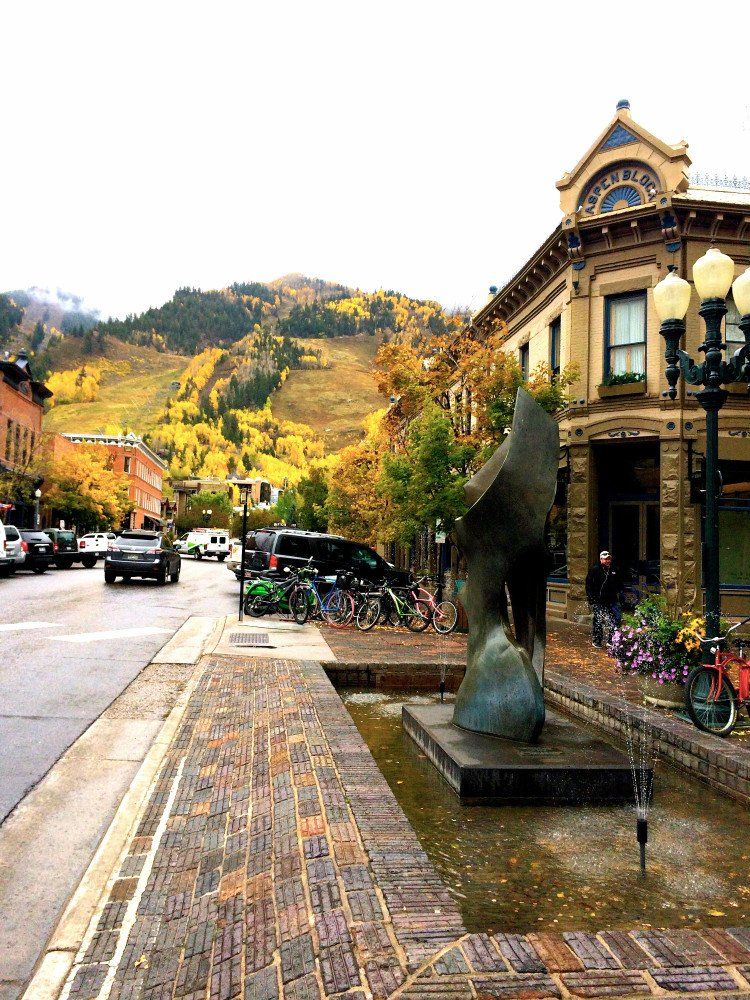 Street scene in Aspen, Colorado with autumn foliage. A sculpture sits in a fountain, with cars, bikes, and buildings lining the street.