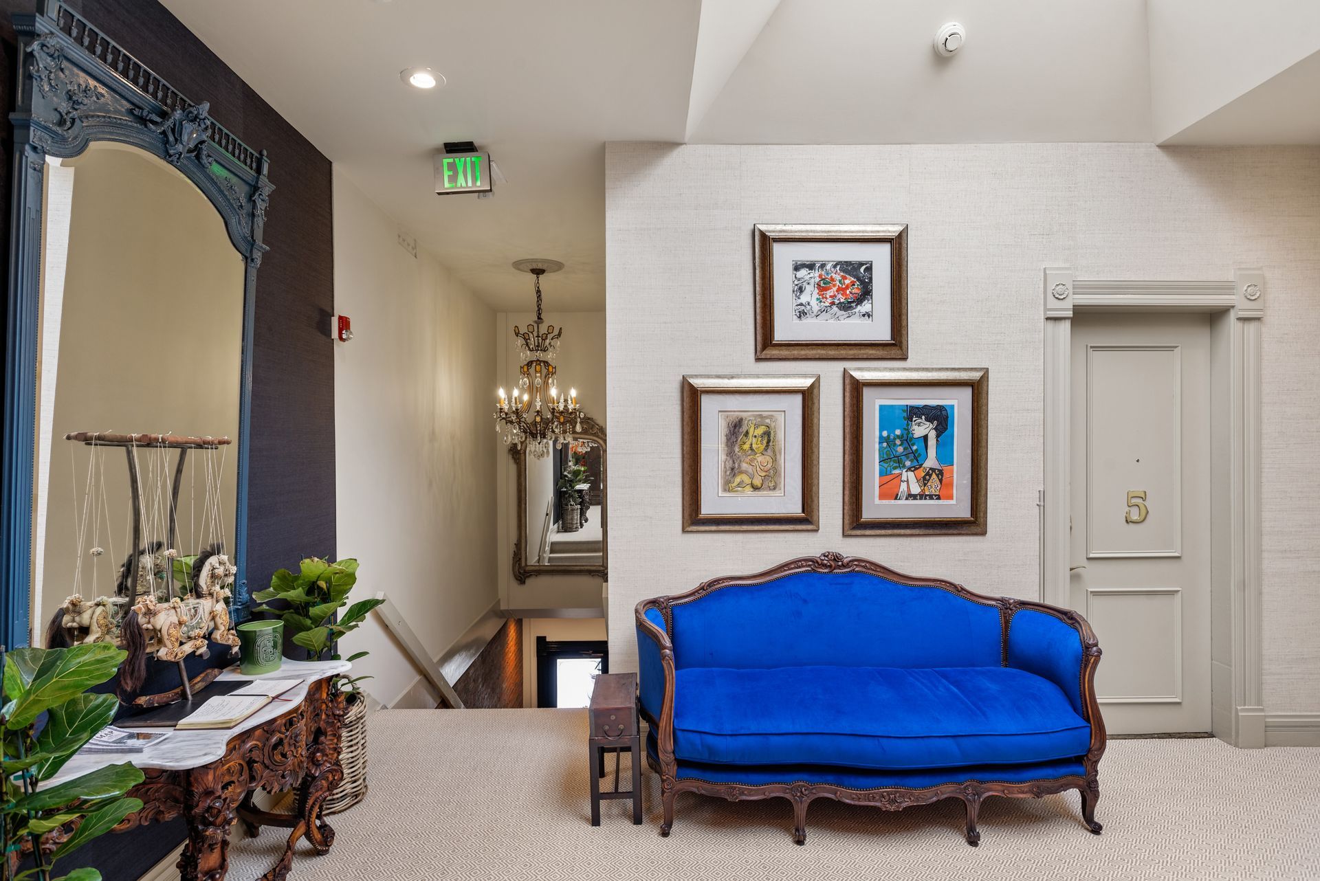 A blue antique couch sits in a hallway with framed art, a large ornate mirror, and a chandelier.