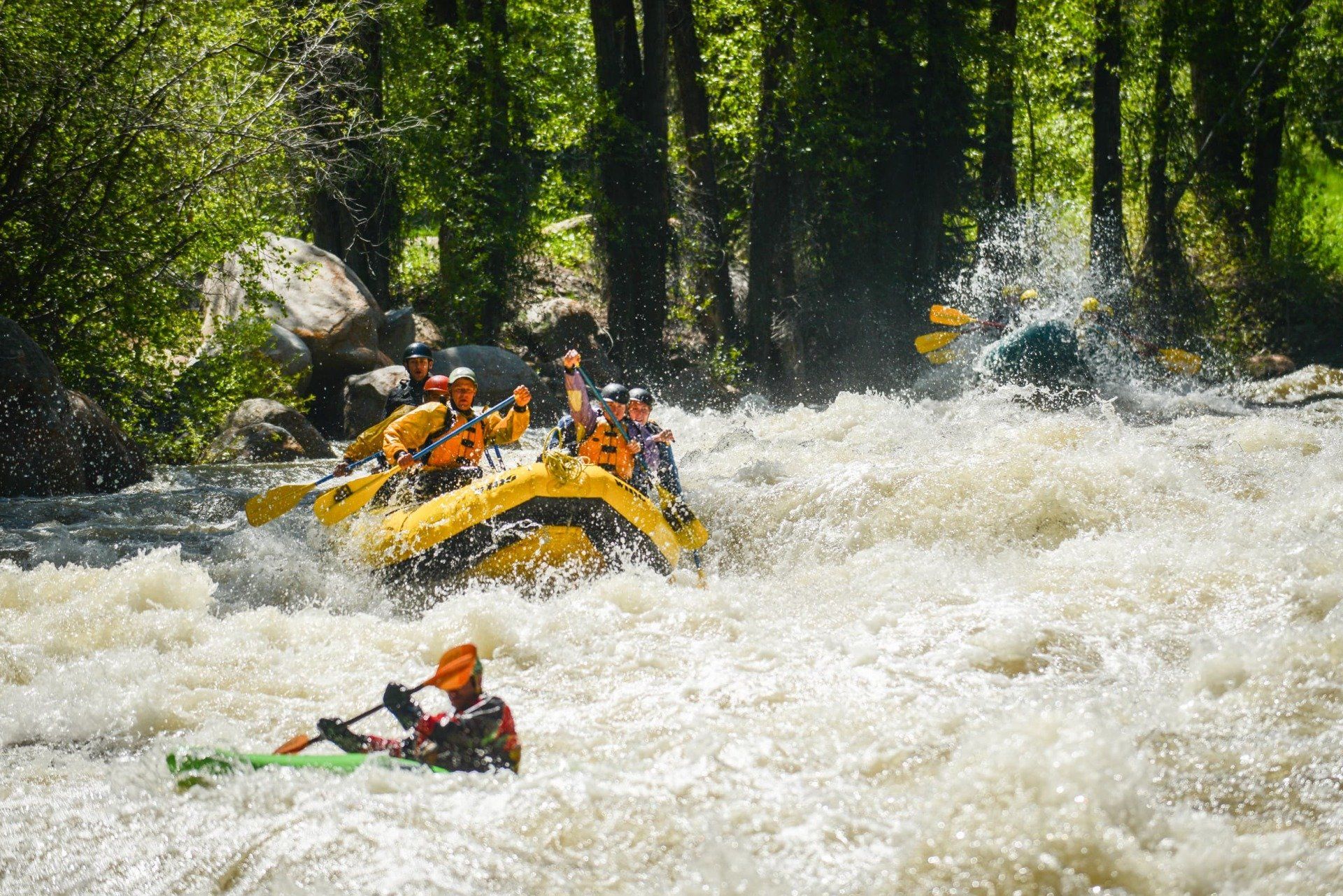 Rafters navigate whitewater rapids in a yellow raft, another person kayaks, trees line the riverbank.