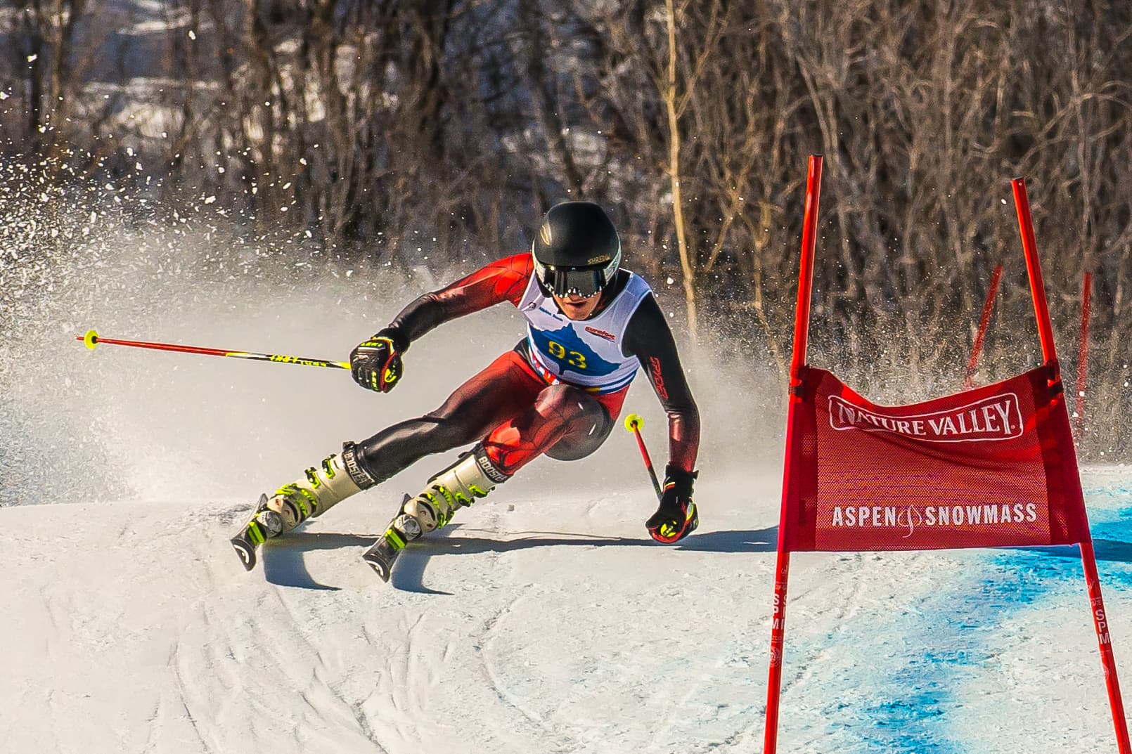 Skier in red and black racing suit, navigating a slalom course, kicking up snow, in front of red flags and a snowy mountain backdrop.