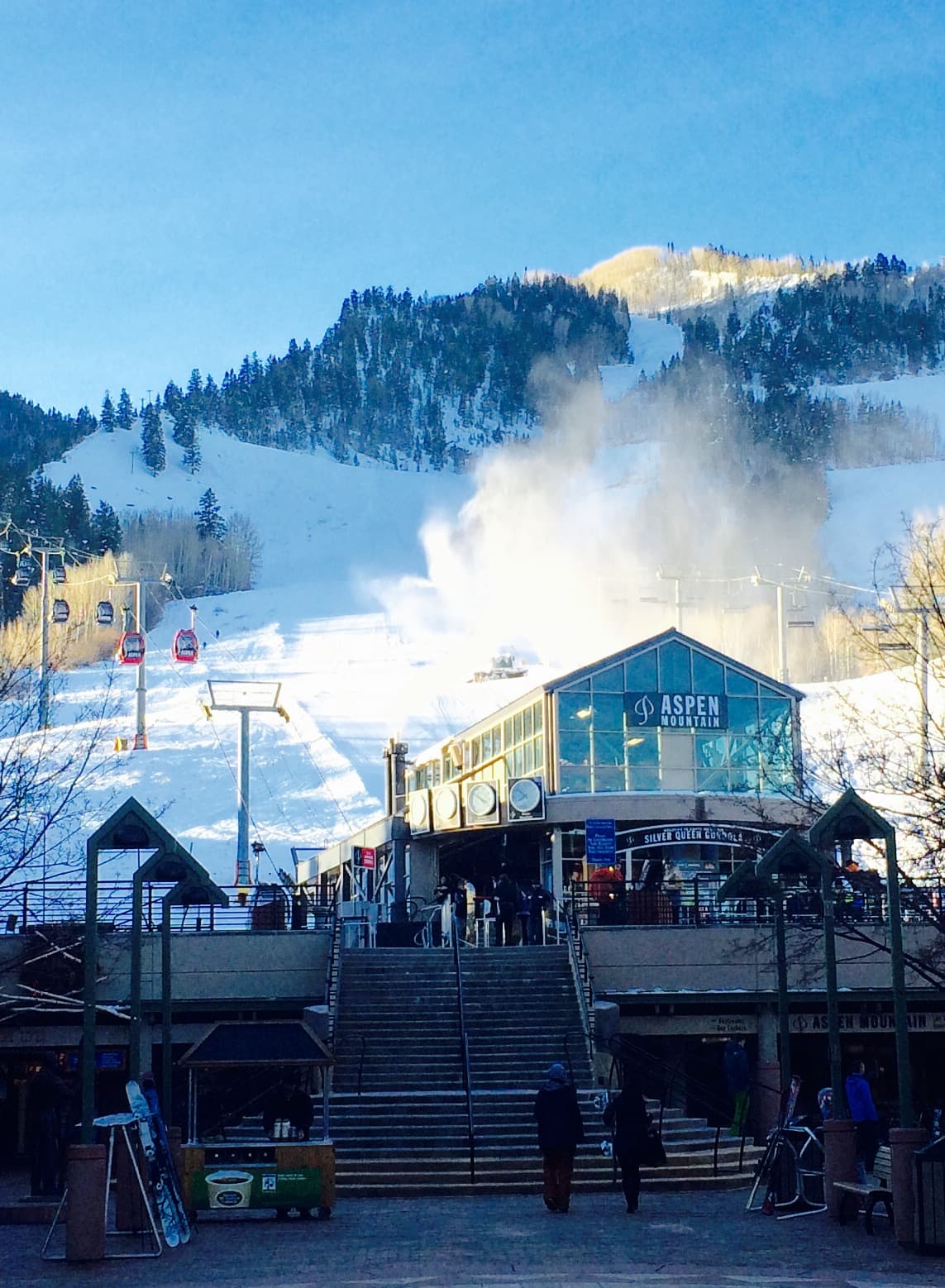 Ski resort on a sunny day. Building with glass front, steps leading up, red ski lift, snowy mountain background.