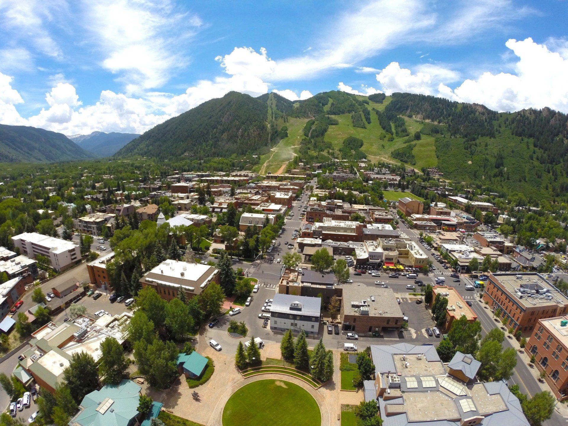 Aerial view of Aspen, Colorado, with buildings, trees, and a mountain in the background on a sunny day.