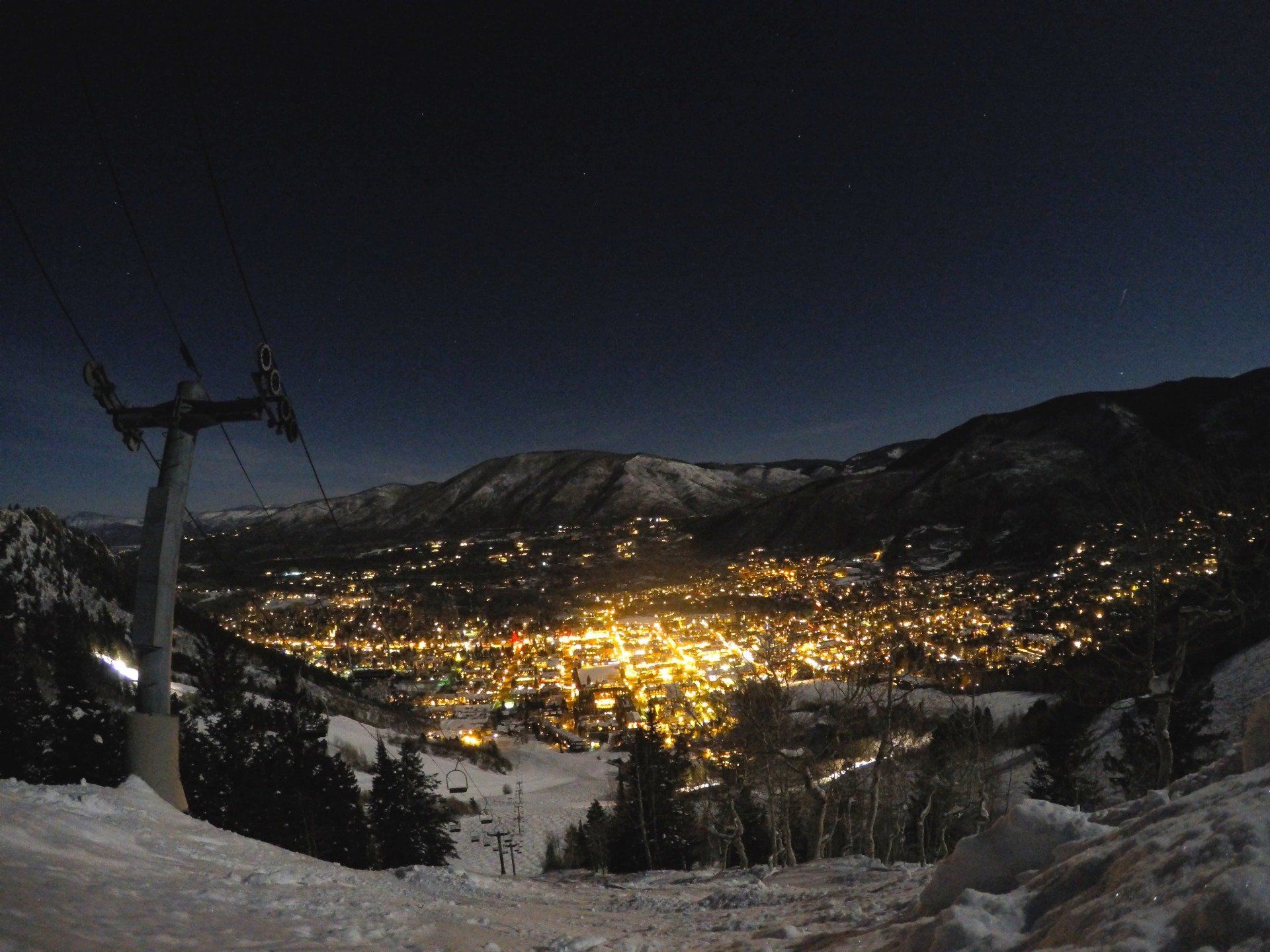 Night view of a snow-covered ski slope overlooking a town lit by golden lights, with a ski lift tower in the foreground.