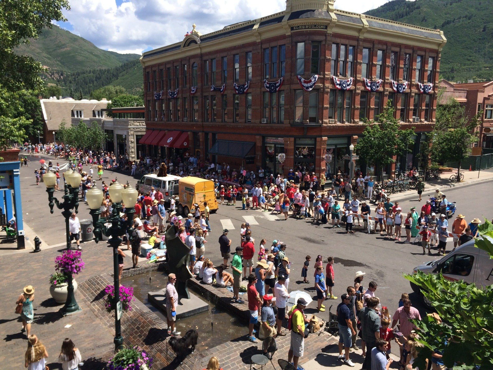 Street scene with a large crowd watching a parade in front of a historic brick building decorated with American flags. Green mountains rise in the background.